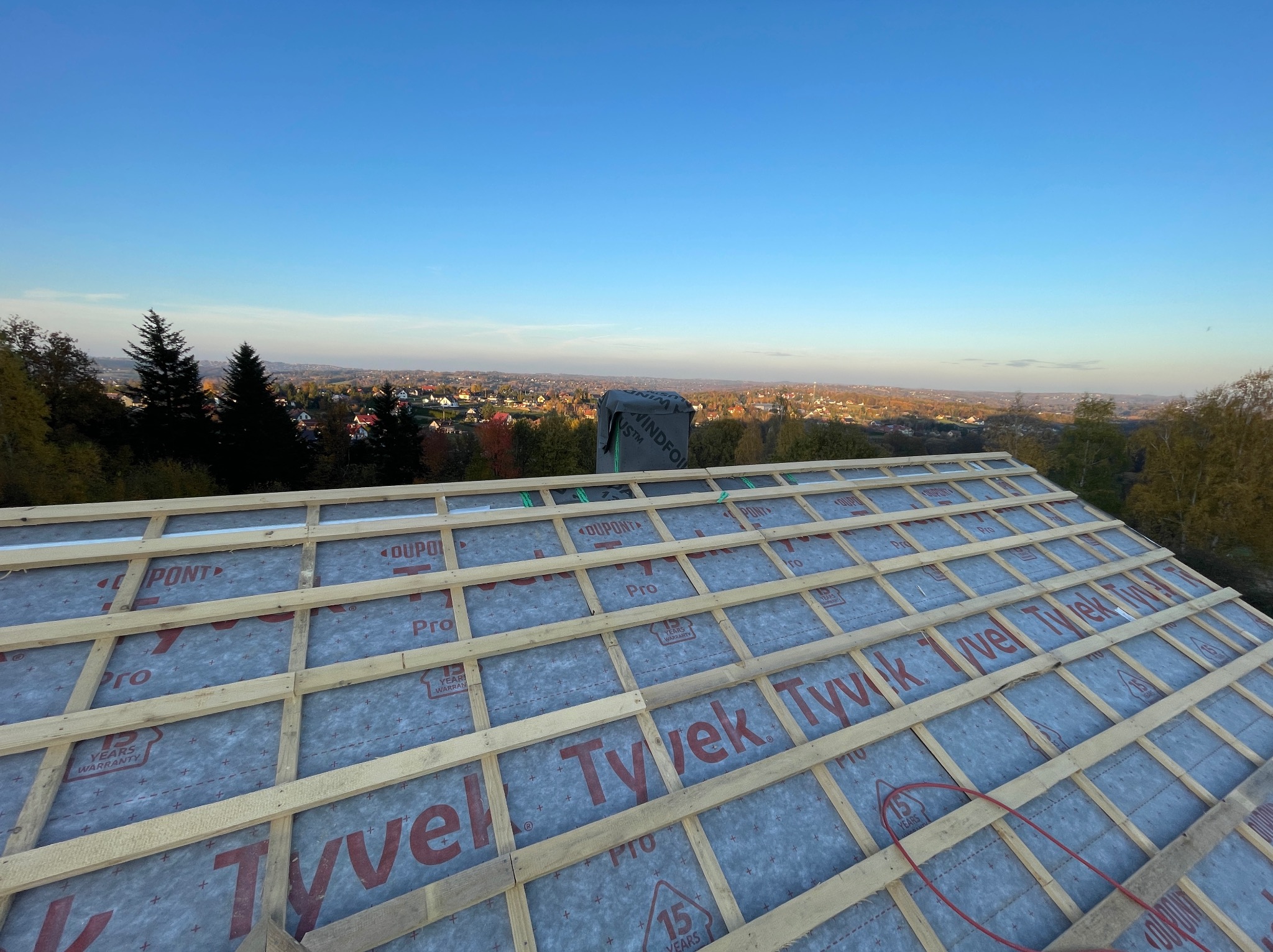 Konstrukcja dachu z membraną Tyvek Pro na tle krajobrazu. Drewniane łaty tworzą regularną siatkę. Widoczne elementy izolacji i komin. Słoneczny dzień, błękitne niebo.