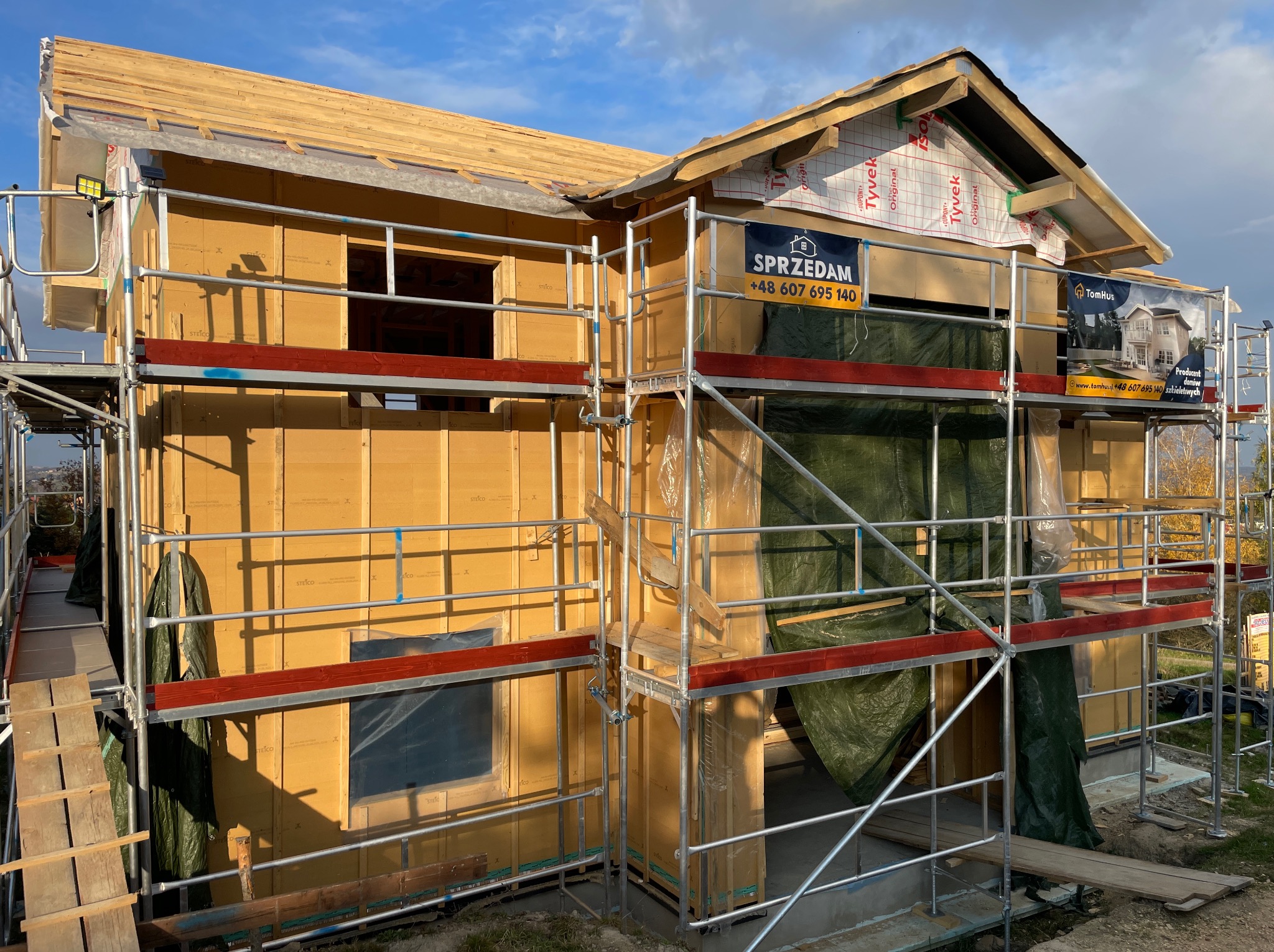 Budowa domu szkieletowego w trakcie: żółte płyty Steico, rusztowania, folia Tyvek. Widoczne reklamy firmy budowlanej i producenta domów szkieletowych TomHus.