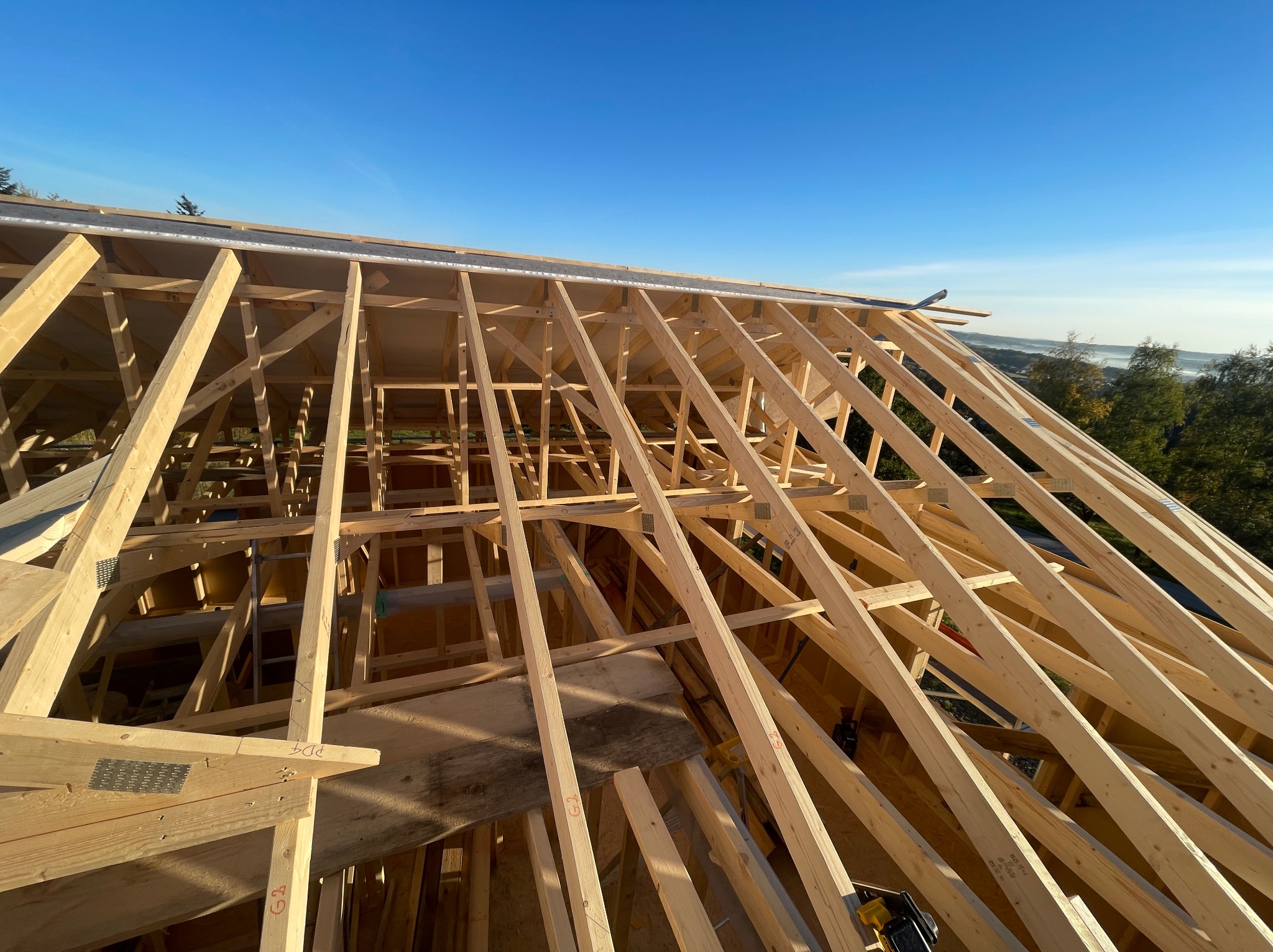 Konstrukcja drewniana dachu w trakcie budowy, widok z góry. Elementy złączone metalowymi płytkami, w tle błękitne niebo i drzewa. Widoczne narzędzia.