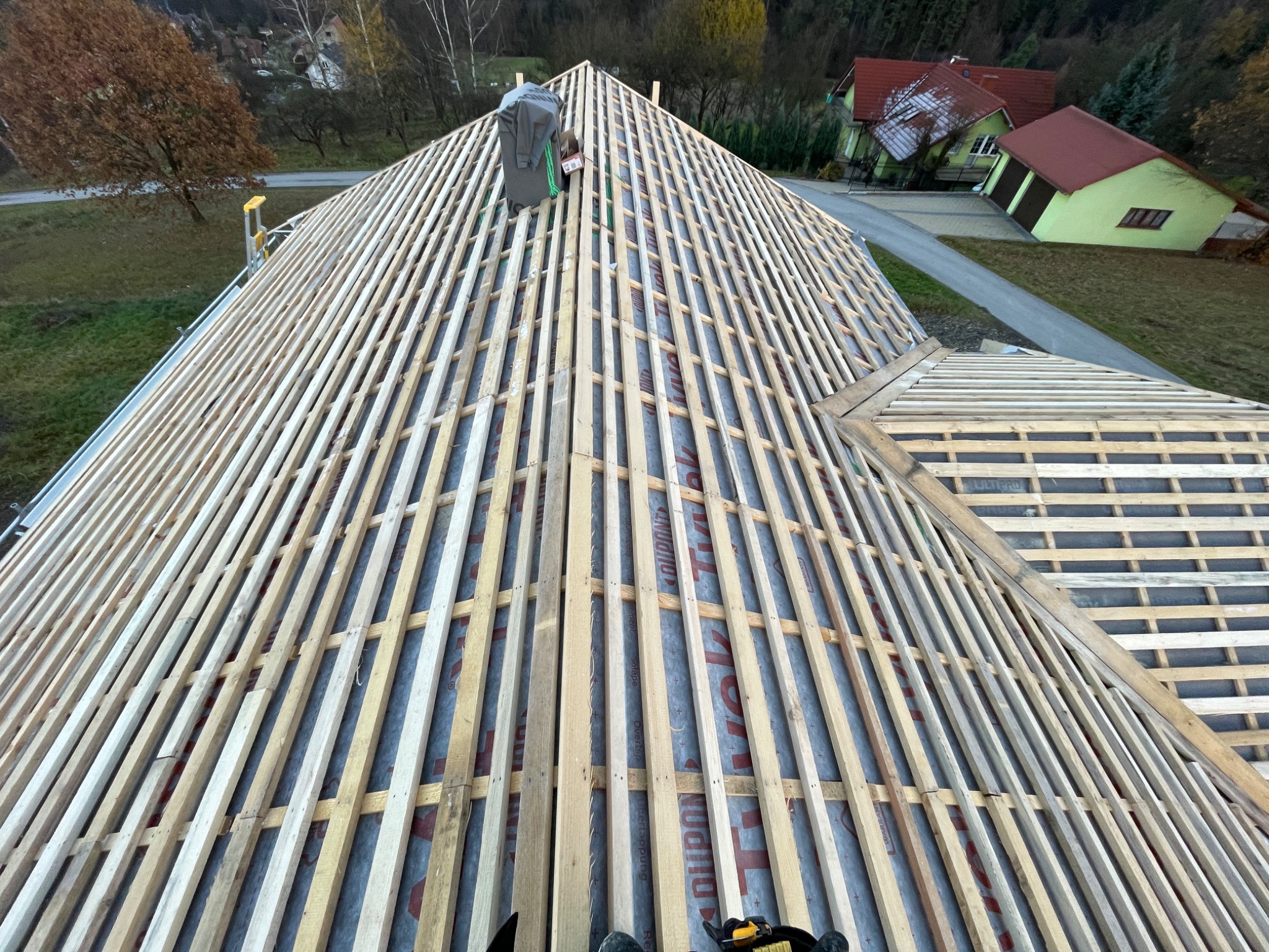 Konstrukcja drewniana dachu z łatami, widok z góry na membranę i elementy mocujące. W tle zabudowa jednorodzinna i drzewa.