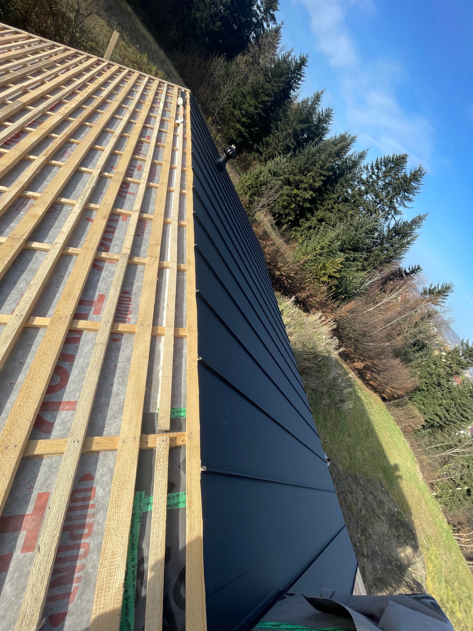 Połowa dachu z czarnym blachy na rąbek, druga połowa w trakcie budowy z drewnianą konstrukcją i folią. W tle drzewa i błękitne niebo.
