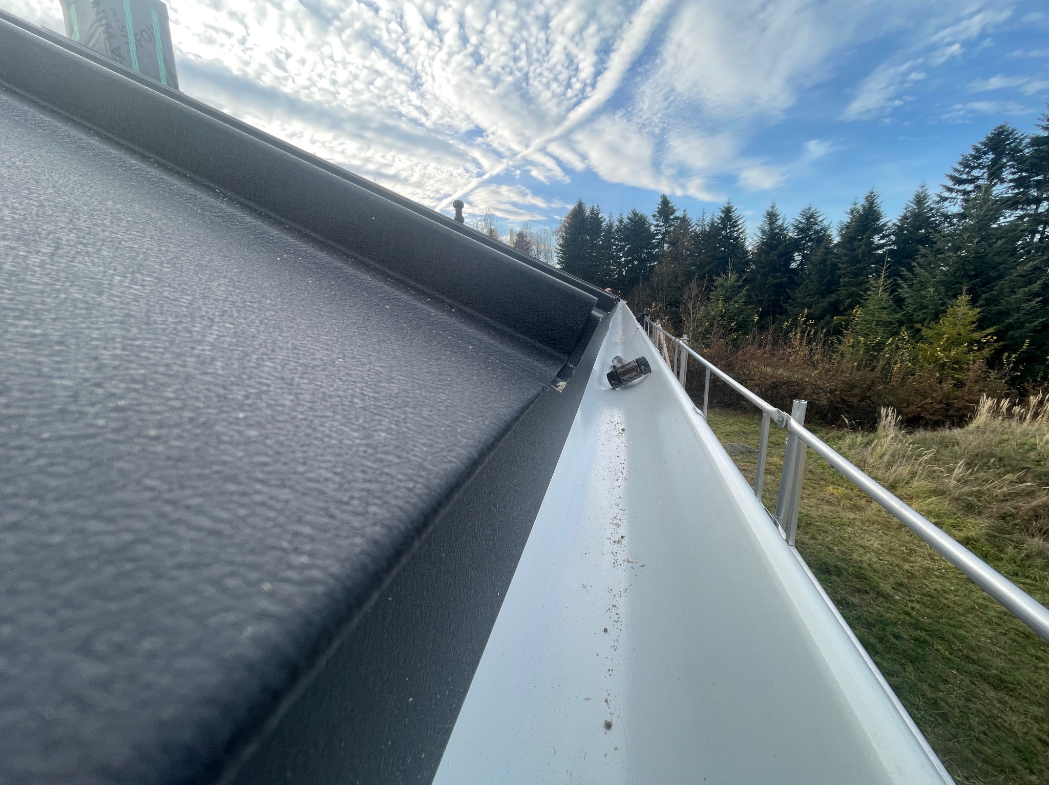 Czarny dach z blachy trapezowej i biała rynna, widok z dołu, na tle błękitnego nieba i drzew. Detal wykończenia dachu.