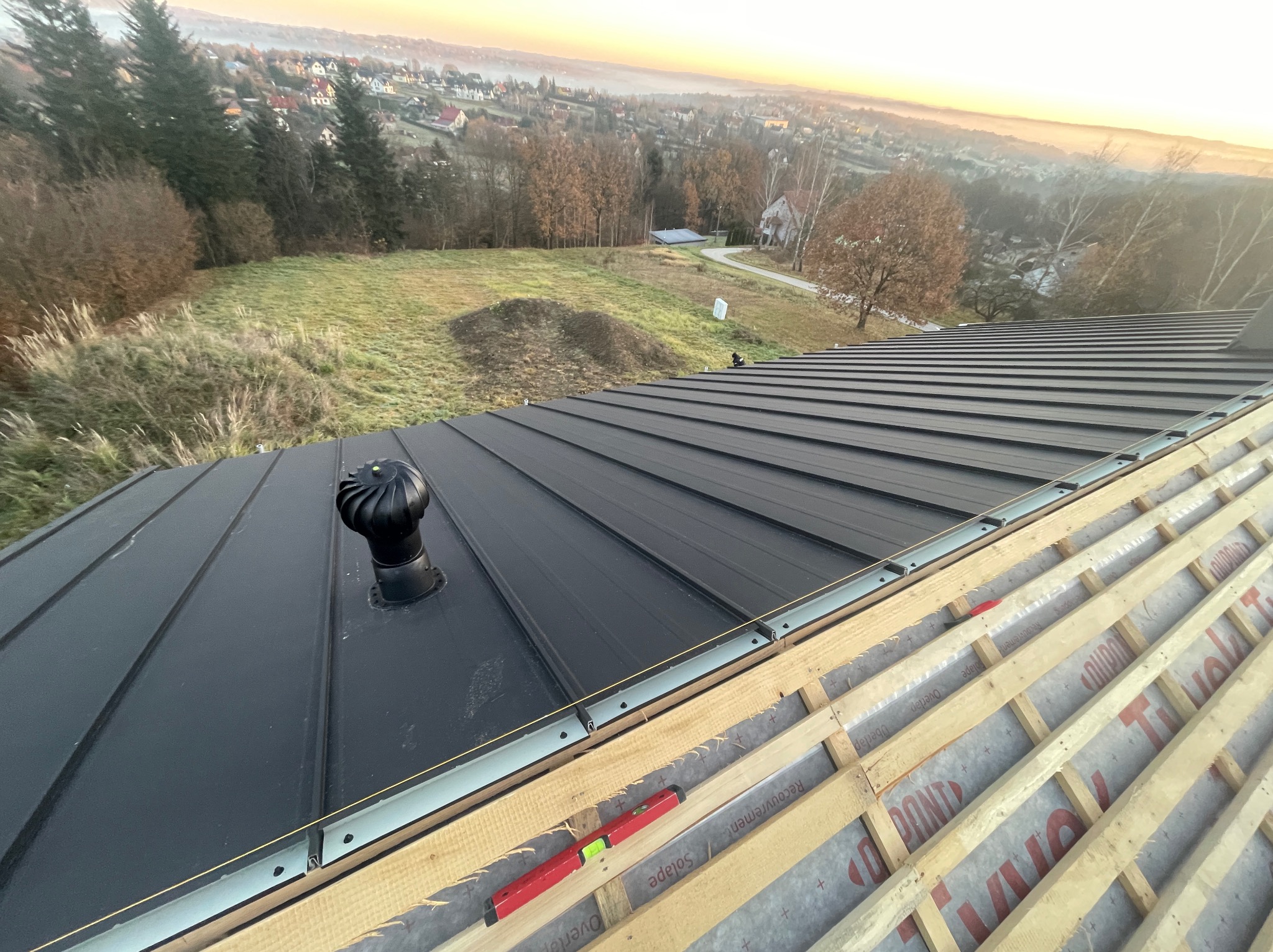 Montaż czarnego dachu panelowego z wentylatorem na domu szkieletowym, widok z góry na konstrukcję i krajobraz w tle o wschodzie słońca.