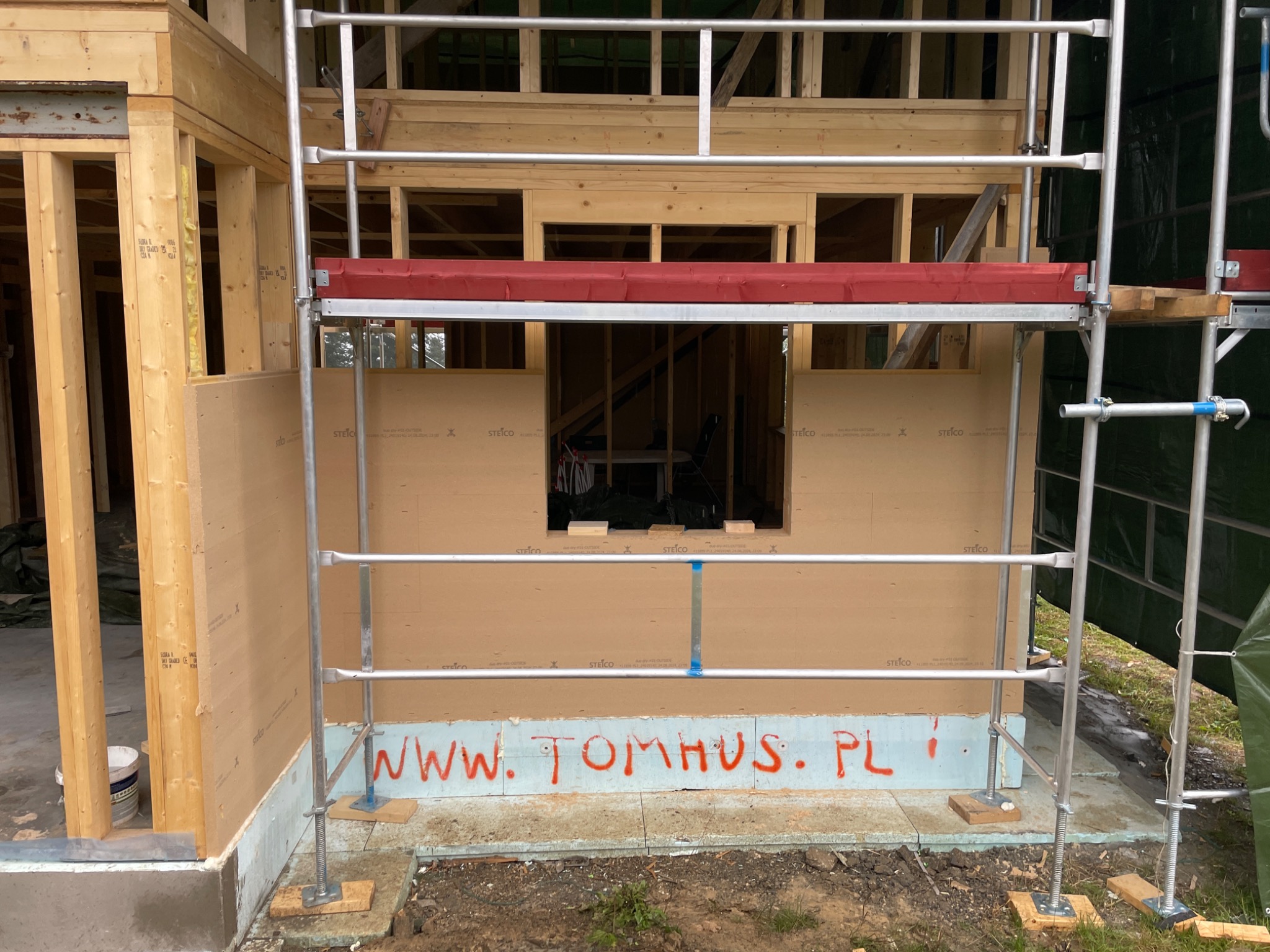 Konstrukcja domu w trakcie budowy, widoczne rusztowanie, drewniane elementy szkieletu i płyty Steico. Na fundamencie napis 'www.tomhus.pl'.