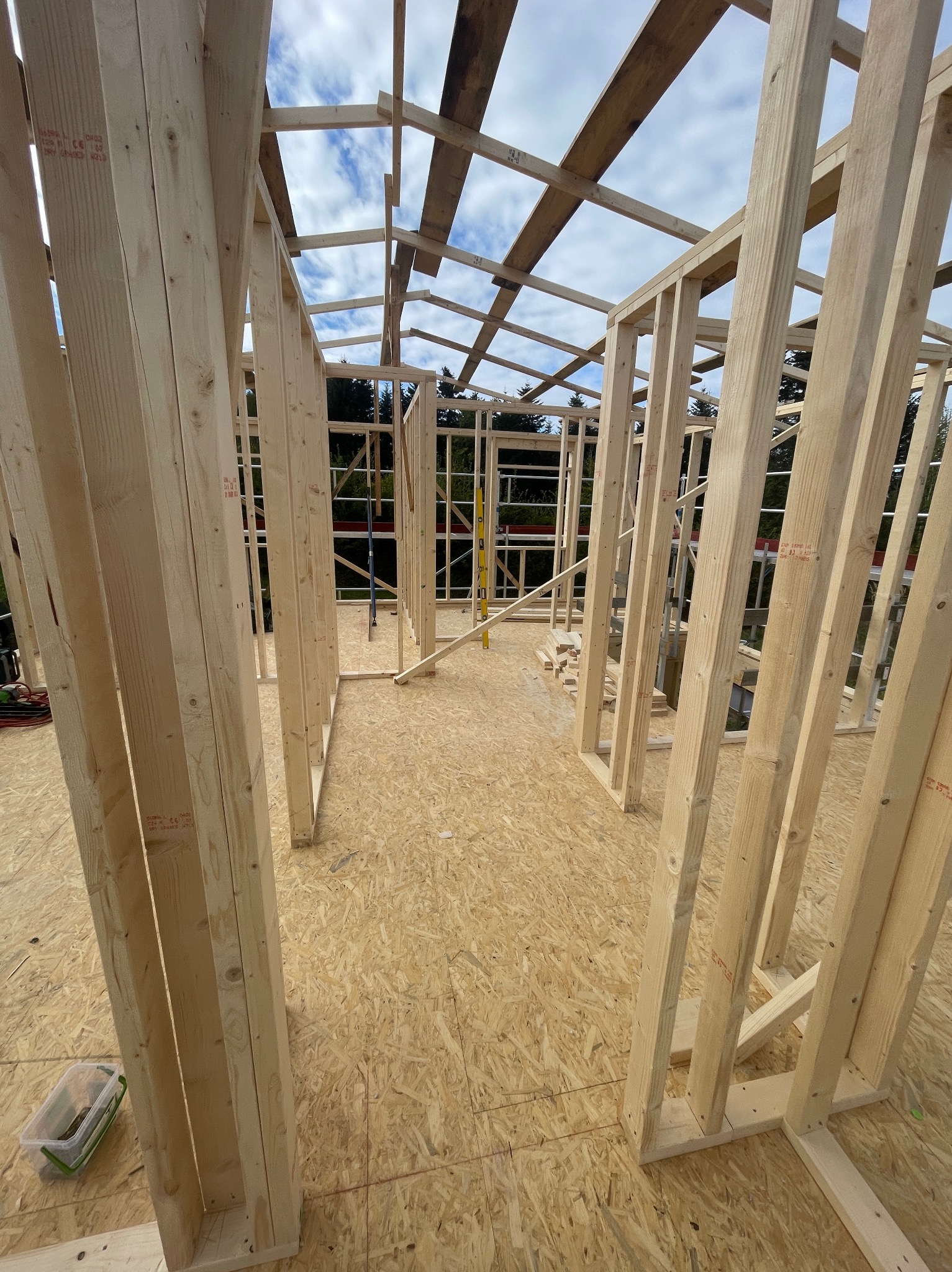 Wnętrze w trakcie budowy: drewniany szkielet domu z widoczną konstrukcją ścian, podłogi z płyt OSB i częściowo zadaszeniem, na tle drzew i błękitnego nieba.