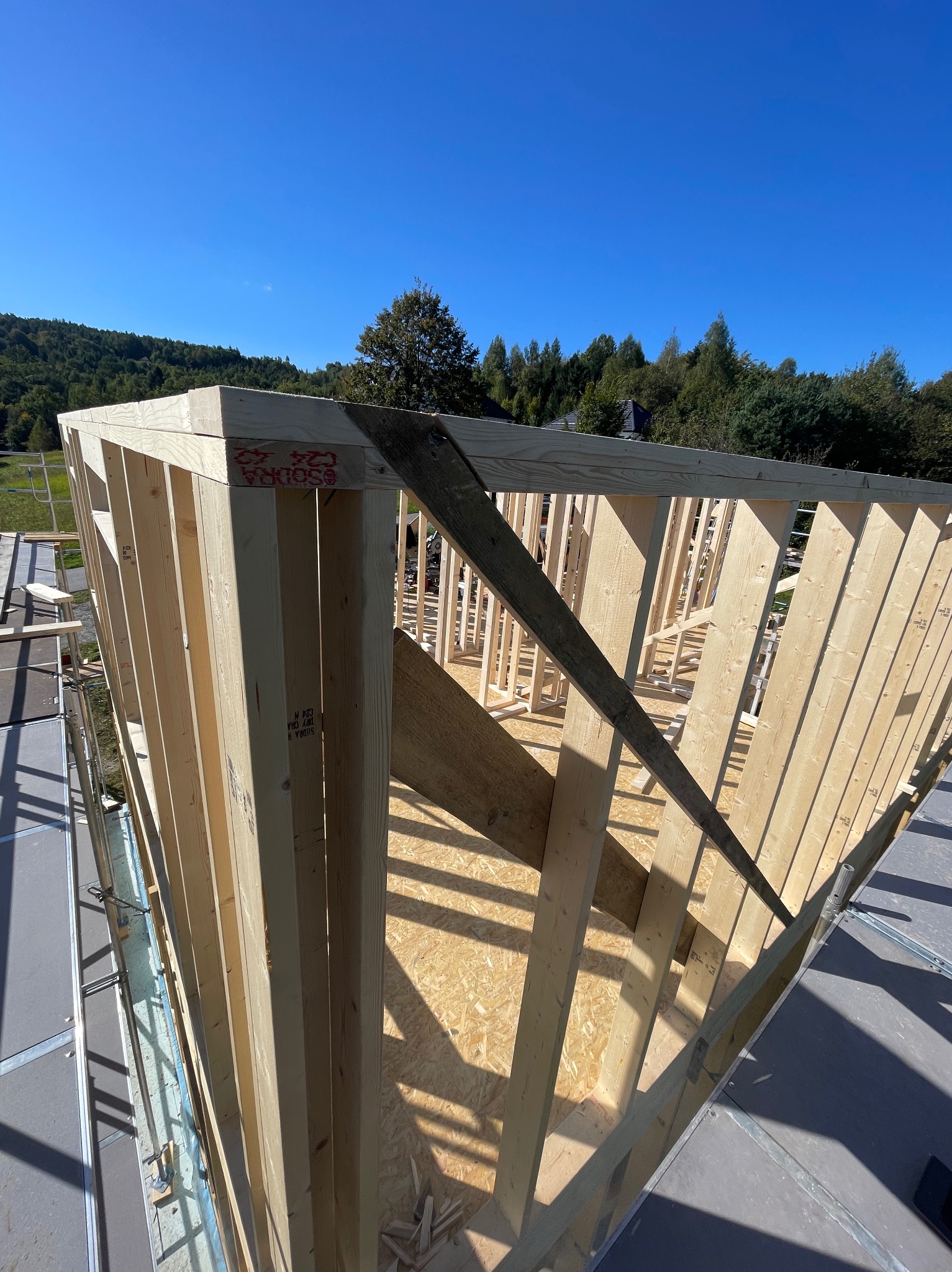 Konstrukcja drewniana domu w trakcie budowy, widok z góry na szkielet ścian i podłogi, z zielonymi wzgórzami i błękitnym niebem w tle.