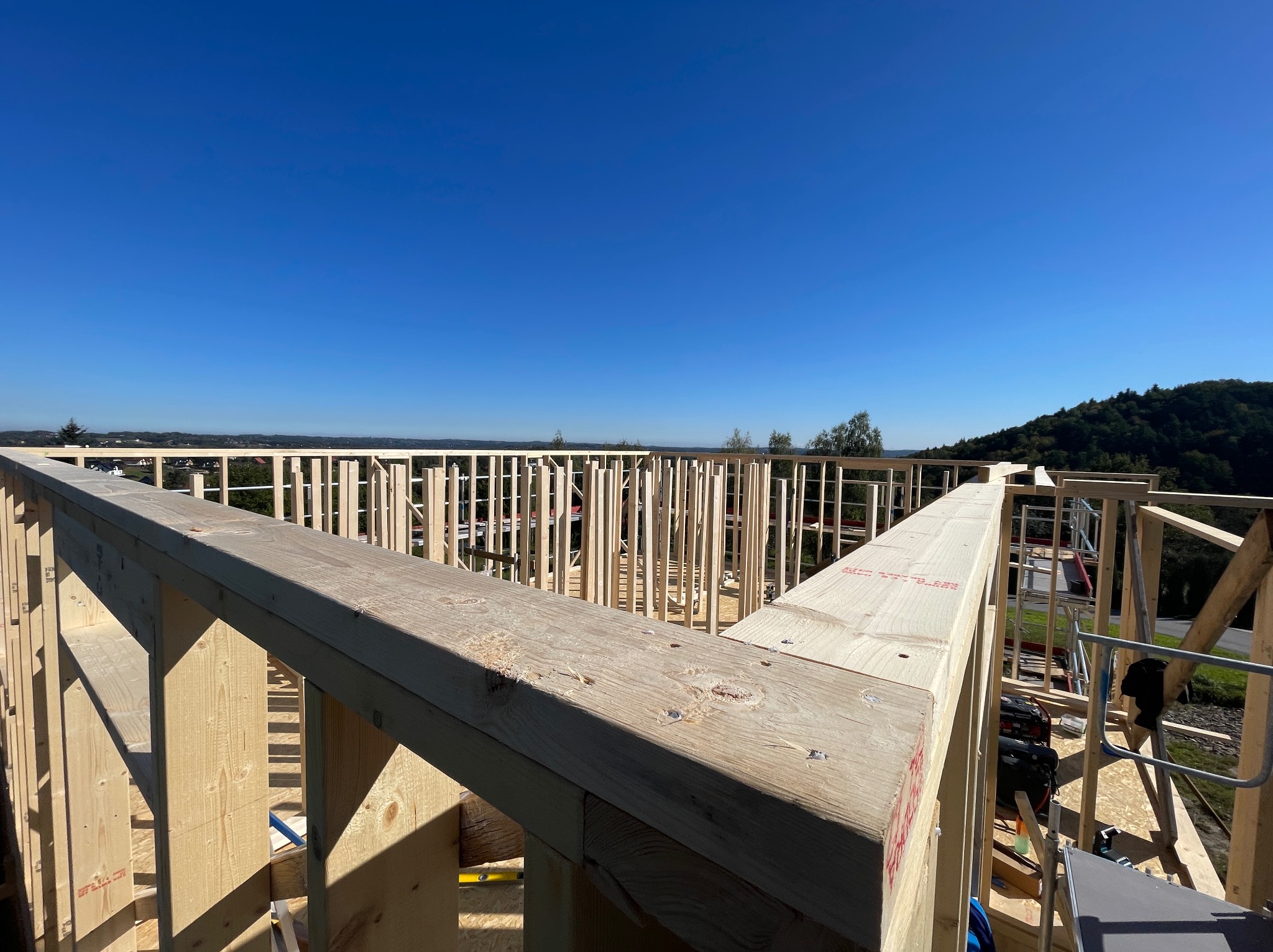 Wnętrze w trakcie budowy: drewniany szkielet domu z widocznymi belkami i słupami, błękitne niebo w tle. Konstrukcja z surowego drewna, w tle zielone wzgórza.