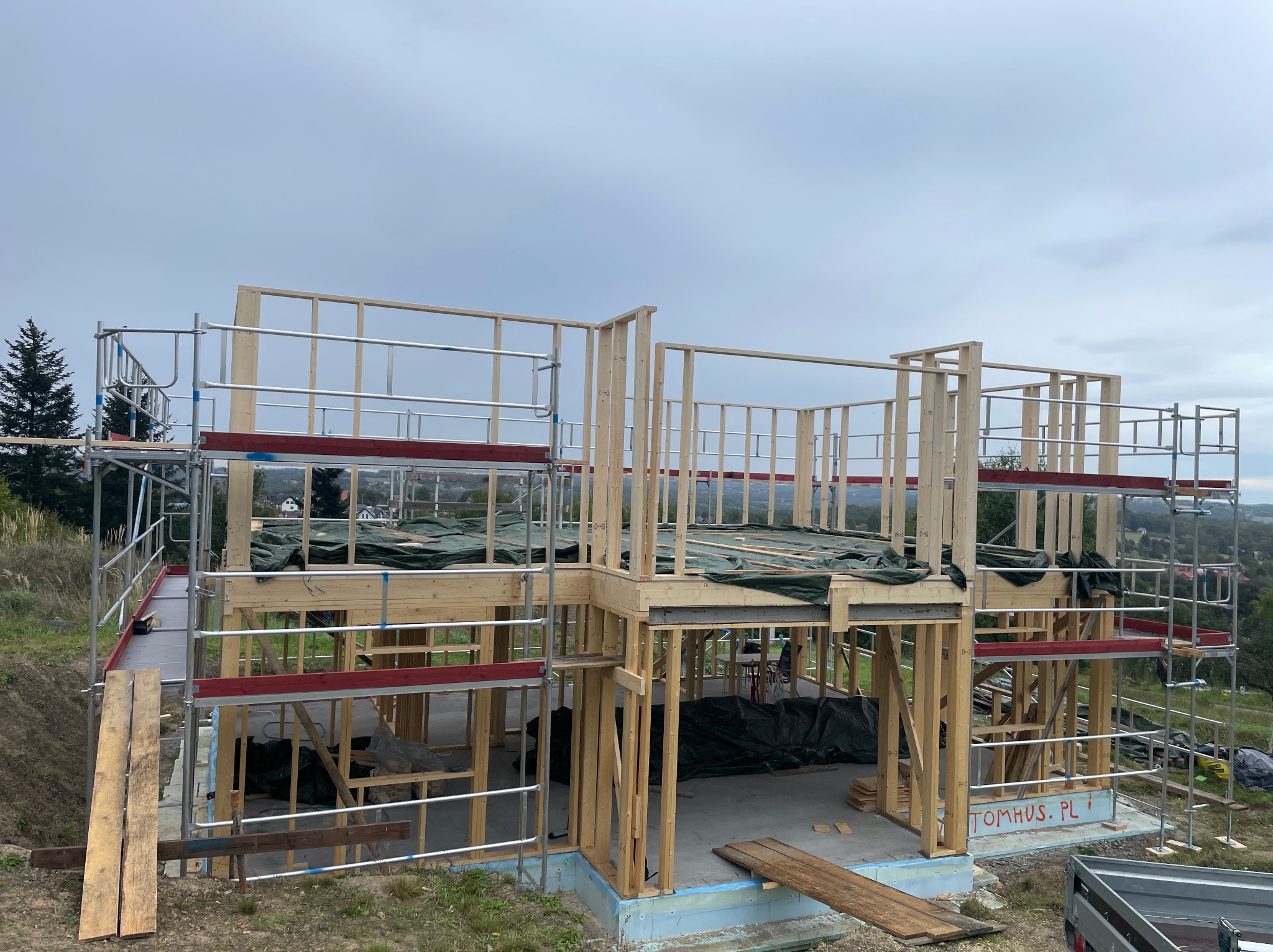 Wznoszenie dwukondygnacyjnego domu w technologii szkieletowej. Widoczne drewniane ramy konstrukcyjne, rusztowania i zabezpieczenia plandekami. Fundamenty z izolacją.