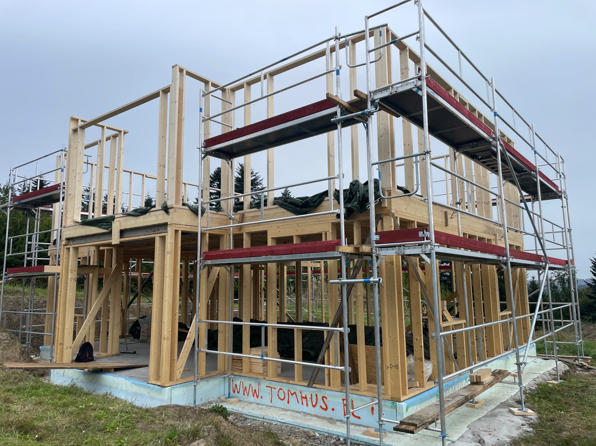 Konstrukcja drewniana domu w trakcie budowy, otoczona rusztowaniami. Widoczny fundament z izolacją oraz częściowo wzniesione ściany szkieletowe. Znak firmowy na fundamencie.