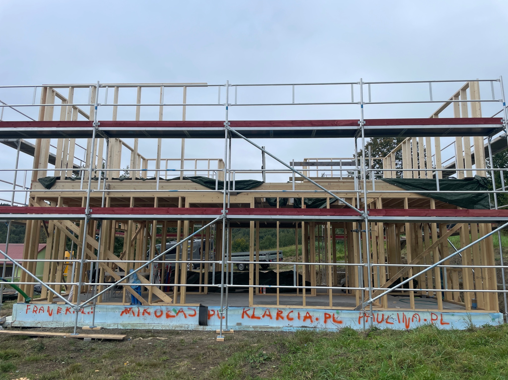 Wznoszenie domu szkieletowego: widok na konstrukcję z rusztowania. Na fundamencie widoczne napisy wykonane czerwoną farbą. W tle drzewa i pochmurne niebo.