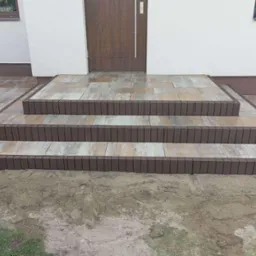 Schody wejściowe z kostki brukowej w odcieniach brązu i beżu, prowadzące do drzwi domu. Widoczne świeżo ułożone obrzeża i fragment chodnika z kostki.