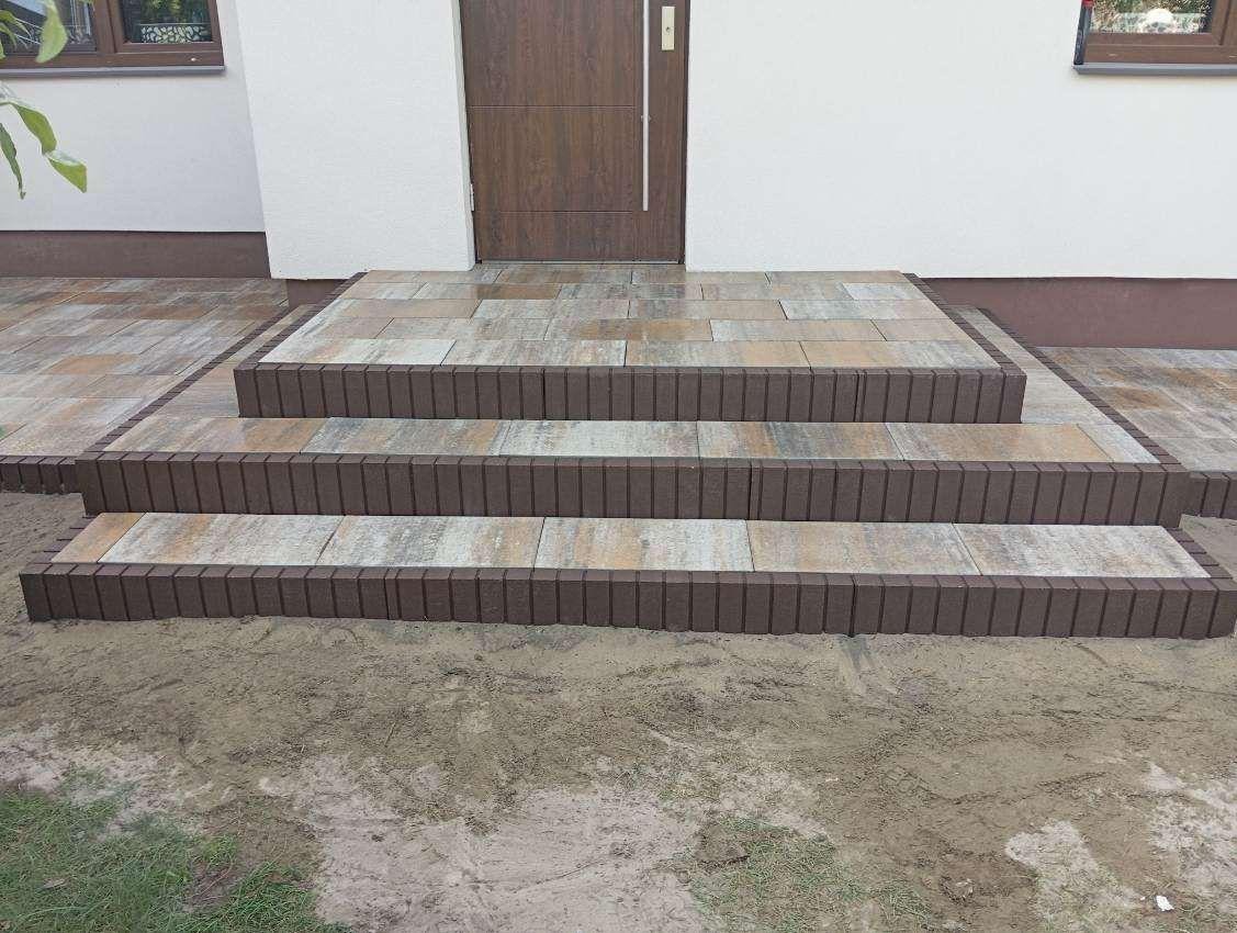 Schody wejściowe z kostki brukowej w odcieniach brązu i beżu, prowadzące do drzwi domu. Widoczne świeżo ułożone obrzeża i fragment chodnika z kostki.