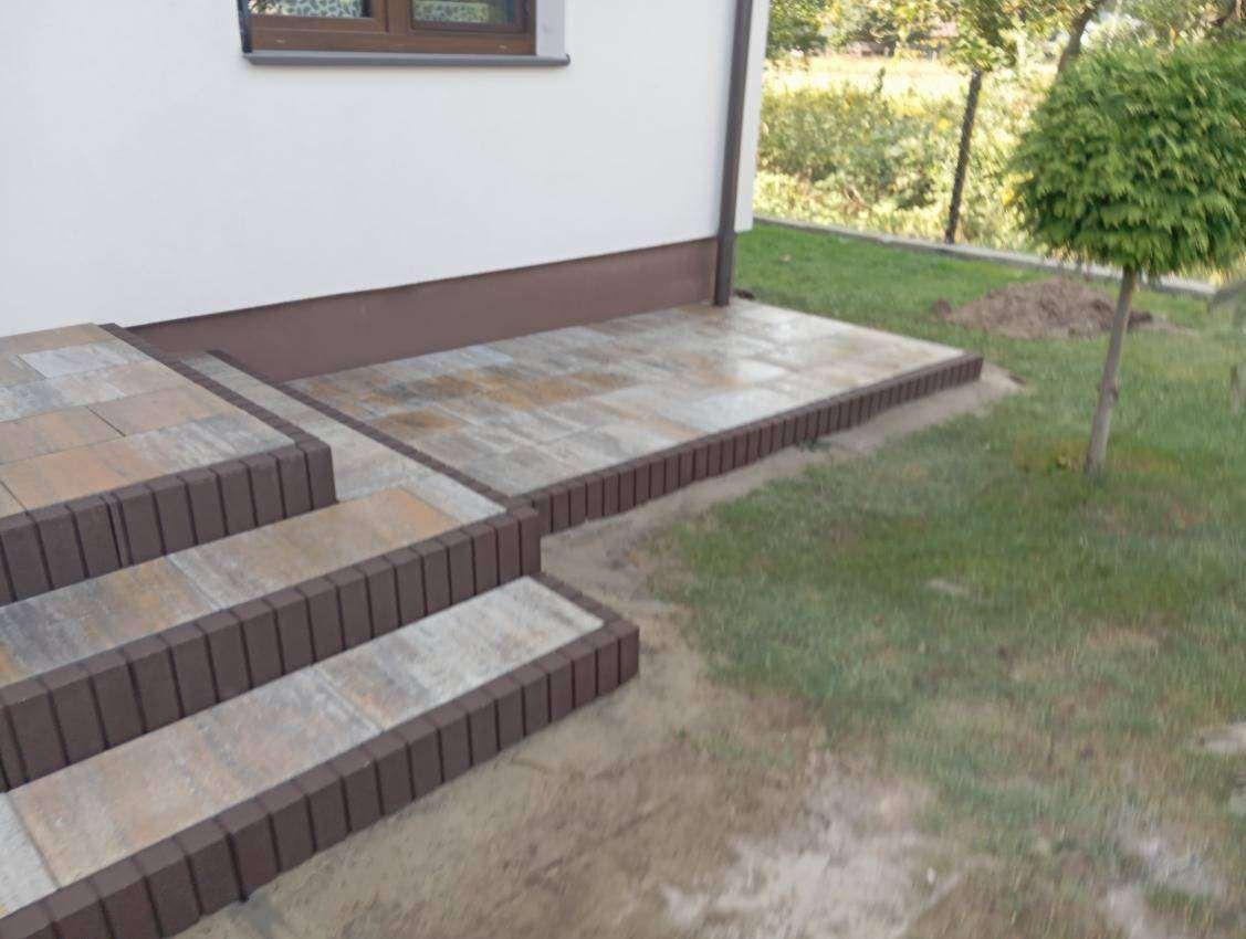 Schody i taras z kostki brukowej w odcieniach szarości i brązu, z obrzeżami, przylegające do białej ściany budynku i trawnika. Widok z bliska.