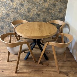 IDEAL FORM Dariusz Kuś - Drewniany okrągły stół z czarną podstawą i czterema krzesłami w jasnym drewnie, na tle wzorzystej tapety i drewnianej podłogi. Nowoczesny design.