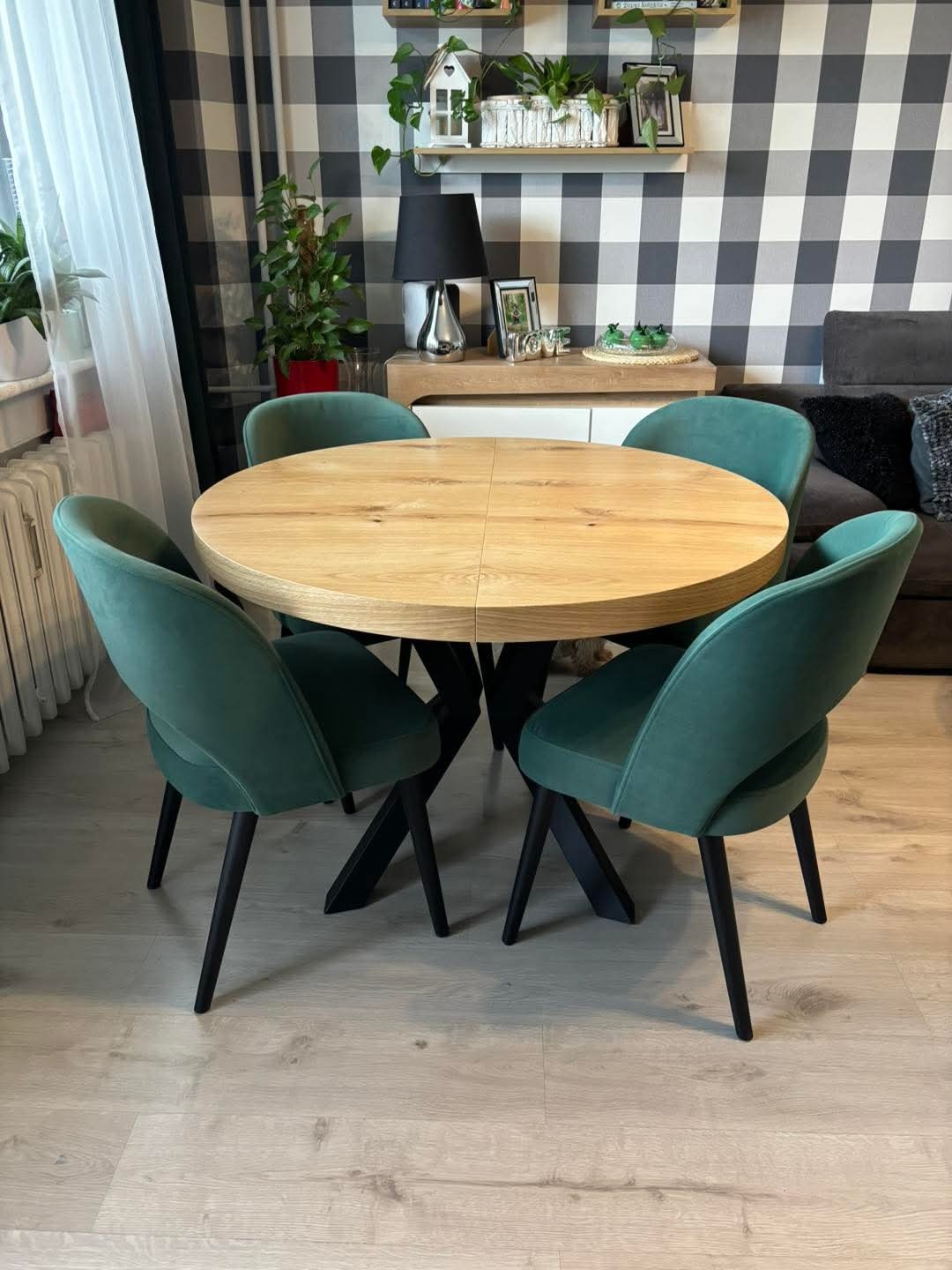 Okrągły, drewniany stół z czterema zielonymi krzesłami w nowoczesnym wnętrzu. W tle tapeta w kratę i rośliny na półkach. Minimalistyczny design.