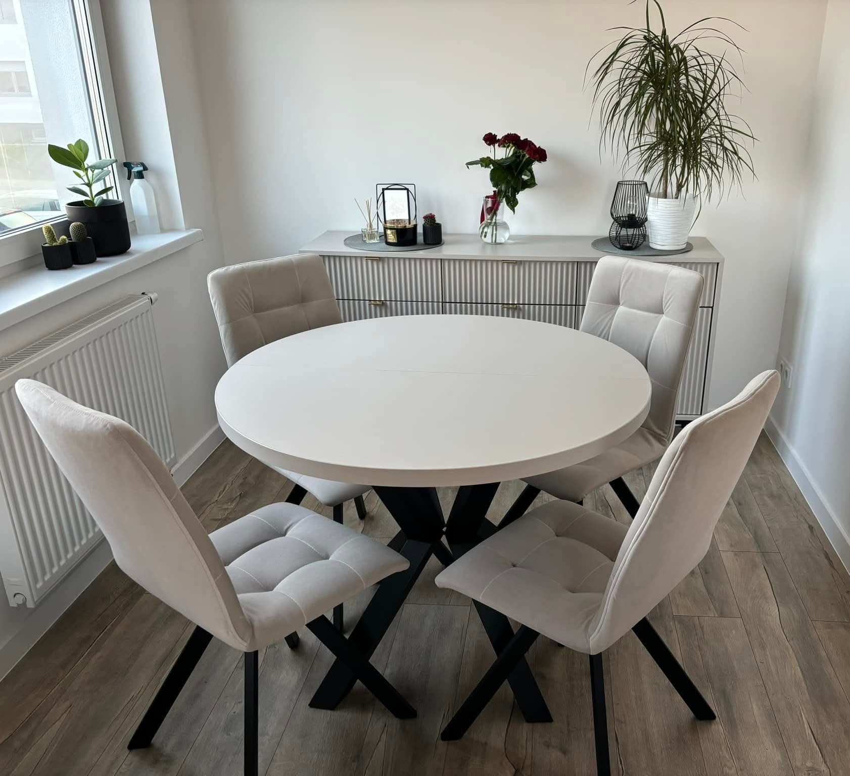 Jadalnia z okrągłym stołem na czarnej podstawie i czterema beżowymi krzesłami. W tle komoda z dekoracjami i kwiatami, całość w jasnej tonacji.