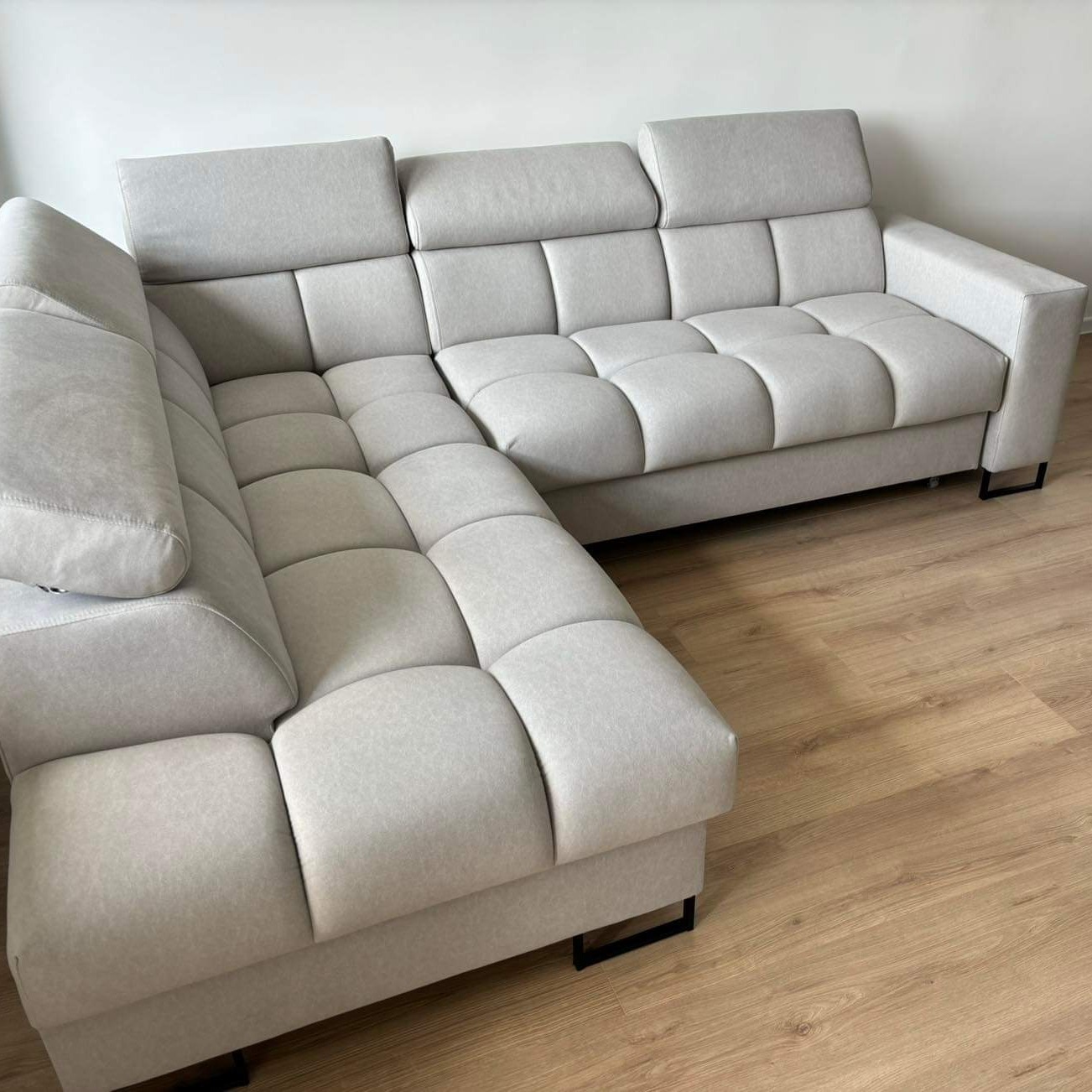 Jasnoszary narożnik z pikowanymi siedziskami i oparciami, na niskich, czarnych nóżkach, ustawiony na drewnianej podłodze, na tle białej ściany.