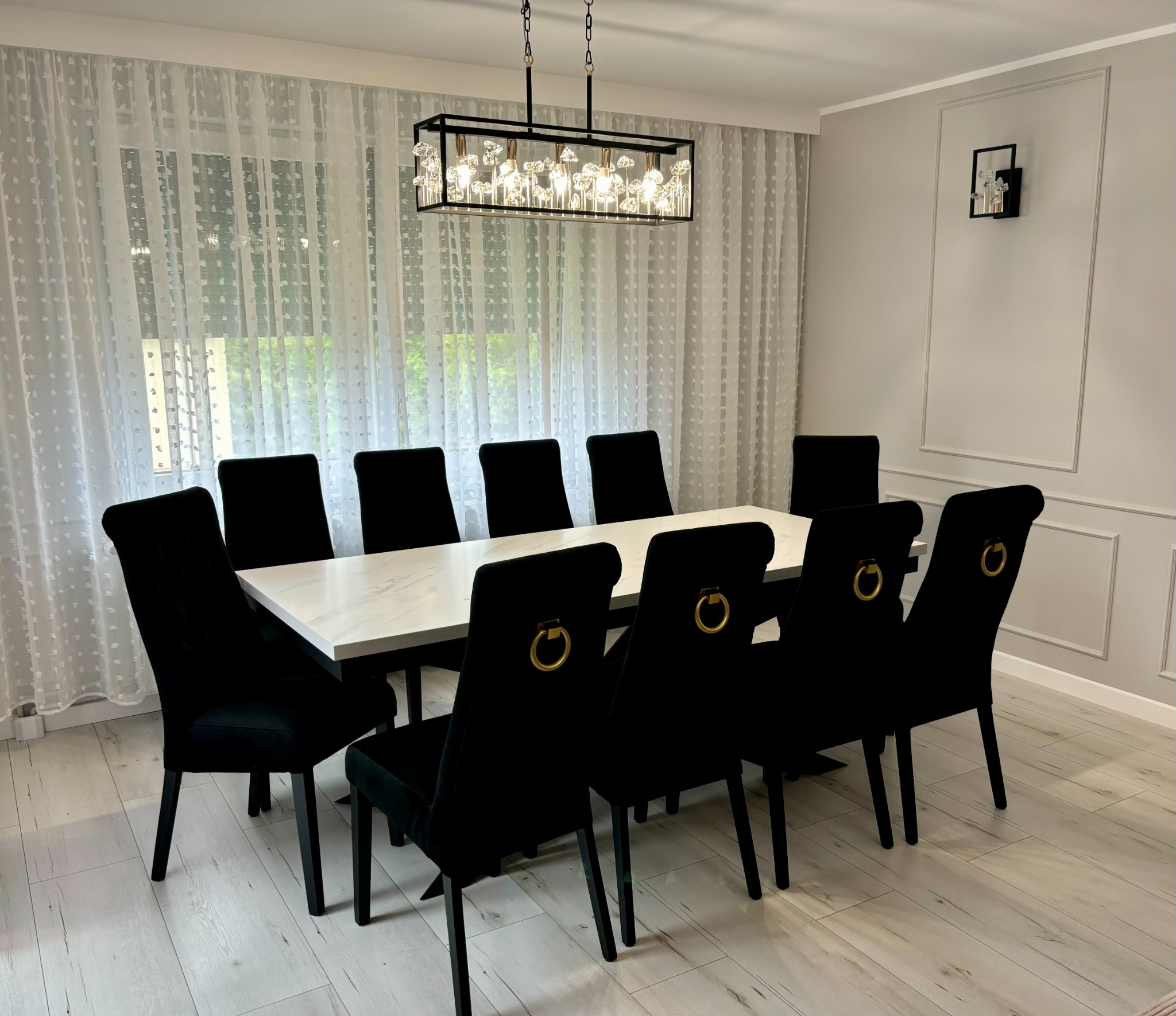 Elegancka jadalnia: stół z marmurowym blatem otoczony czarnymi krzesłami z ozdobnymi złotymi pierścieniami. Nad stołem designerska lampa. W tle okno z białymi firanami.