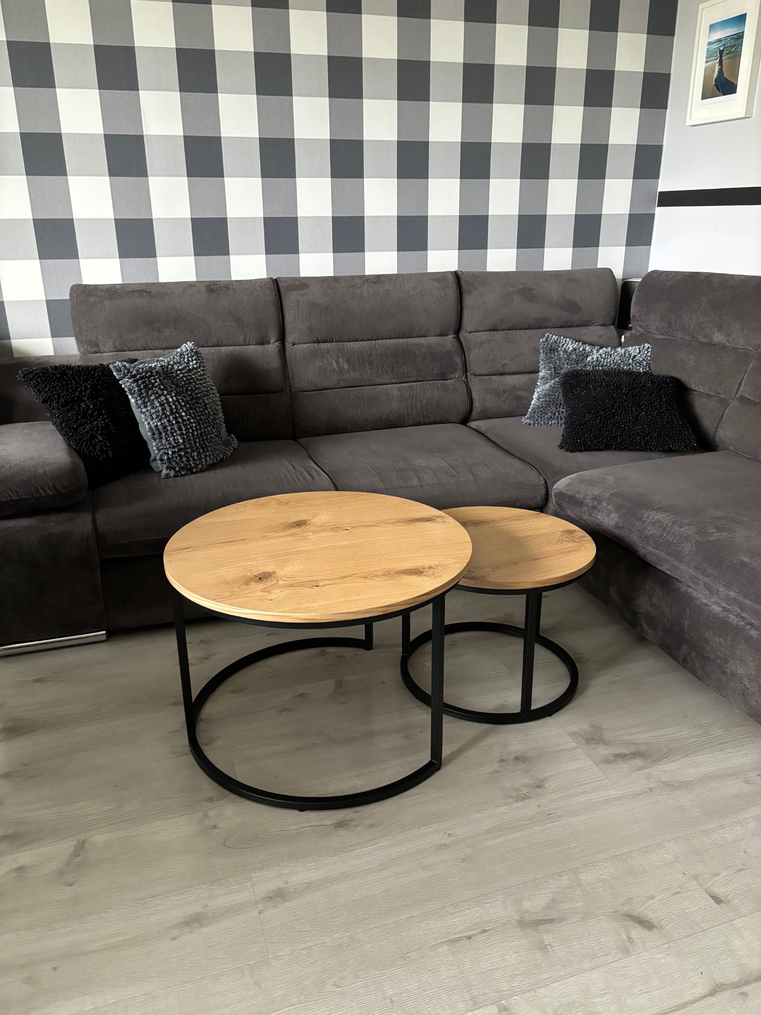 Dwa okrągłe stoliki kawowe z drewnianymi blatami i czarnymi metalowymi nogami na tle szarej sofy i wzorzystej tapety w kratę.