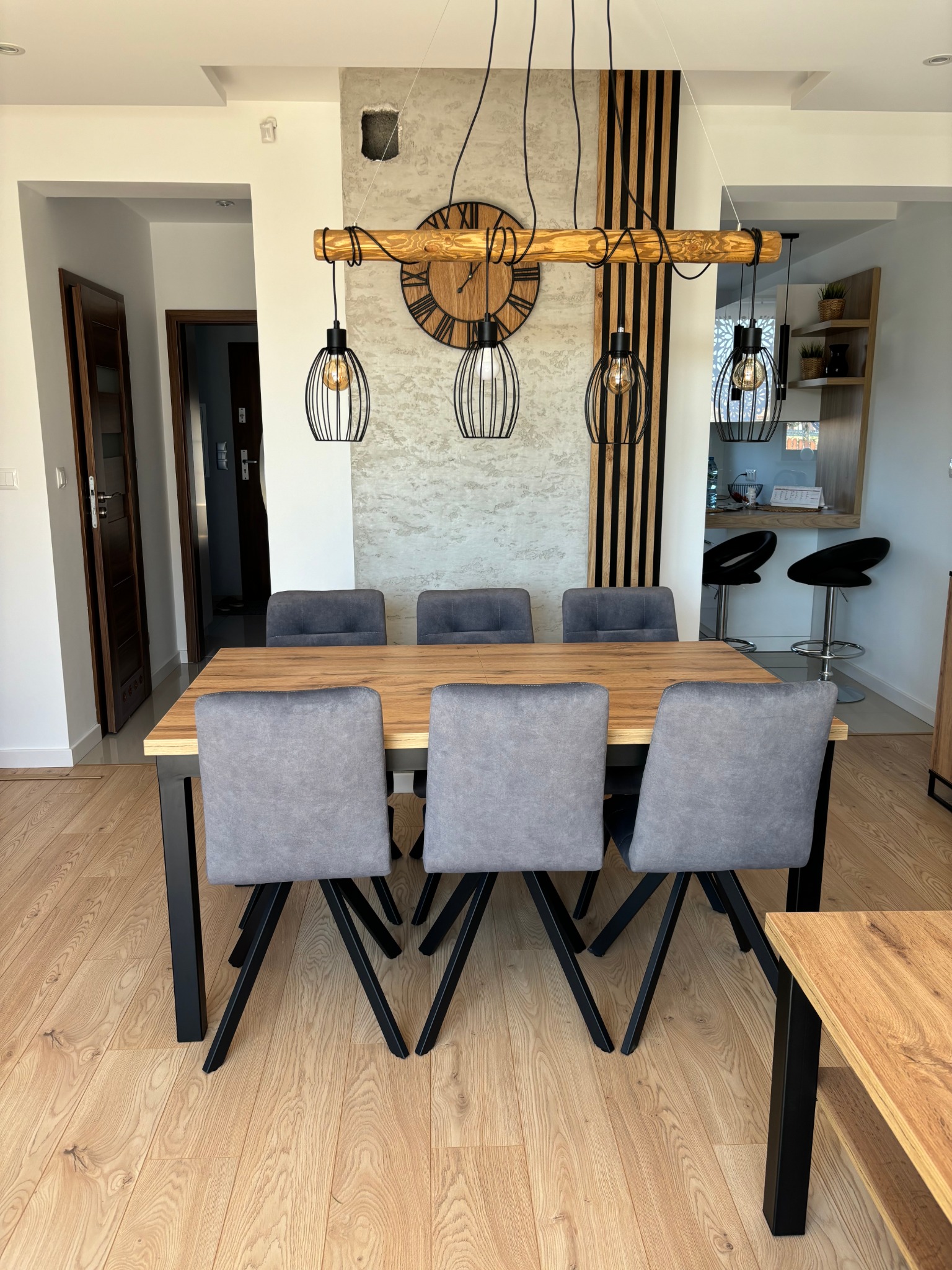 Jadalnia w stylu loft z drewnianym stołem, szarymi krzesłami i dekoracyjnym oświetleniem. Na ścianie betonowa faktura i drewniany zegar.