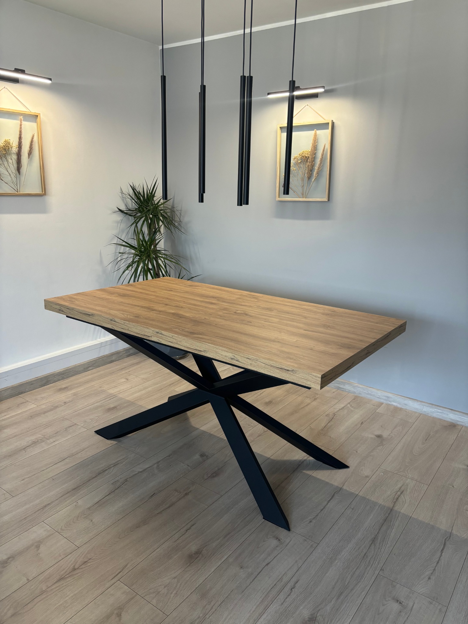 Stół z drewnianym blatem i czarną, geometryczną podstawą w nowoczesnym wnętrzu z dekoracjami ściennymi i lampami. Minimalistyczny design, jasne drewno.