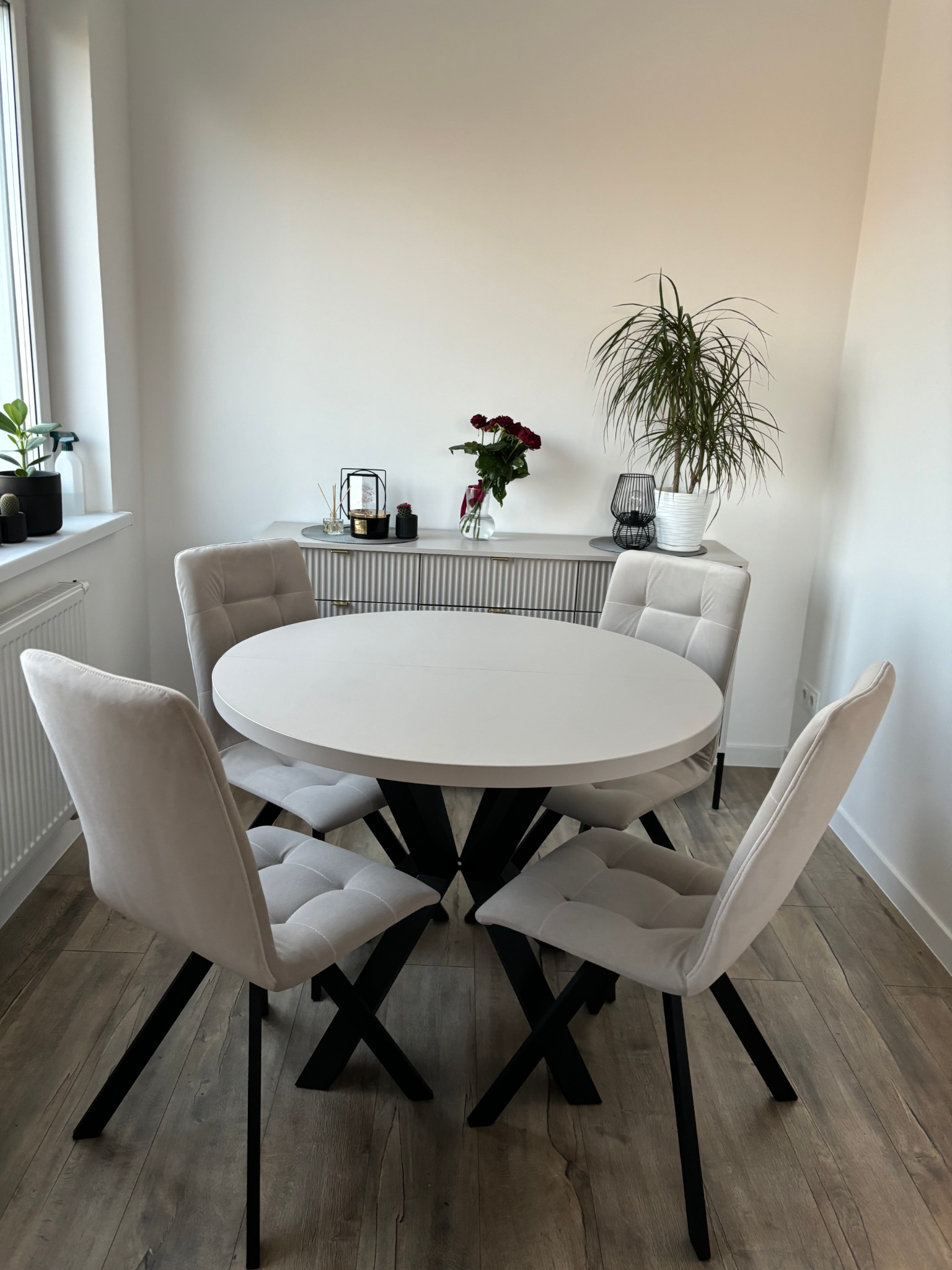 Jasny stół z krzesłami na tle komody z dekoracjami i roślinami. Minimalistyczne wnętrze w stonowanych barwach, z drewnianą podłogą i dużym oknem.