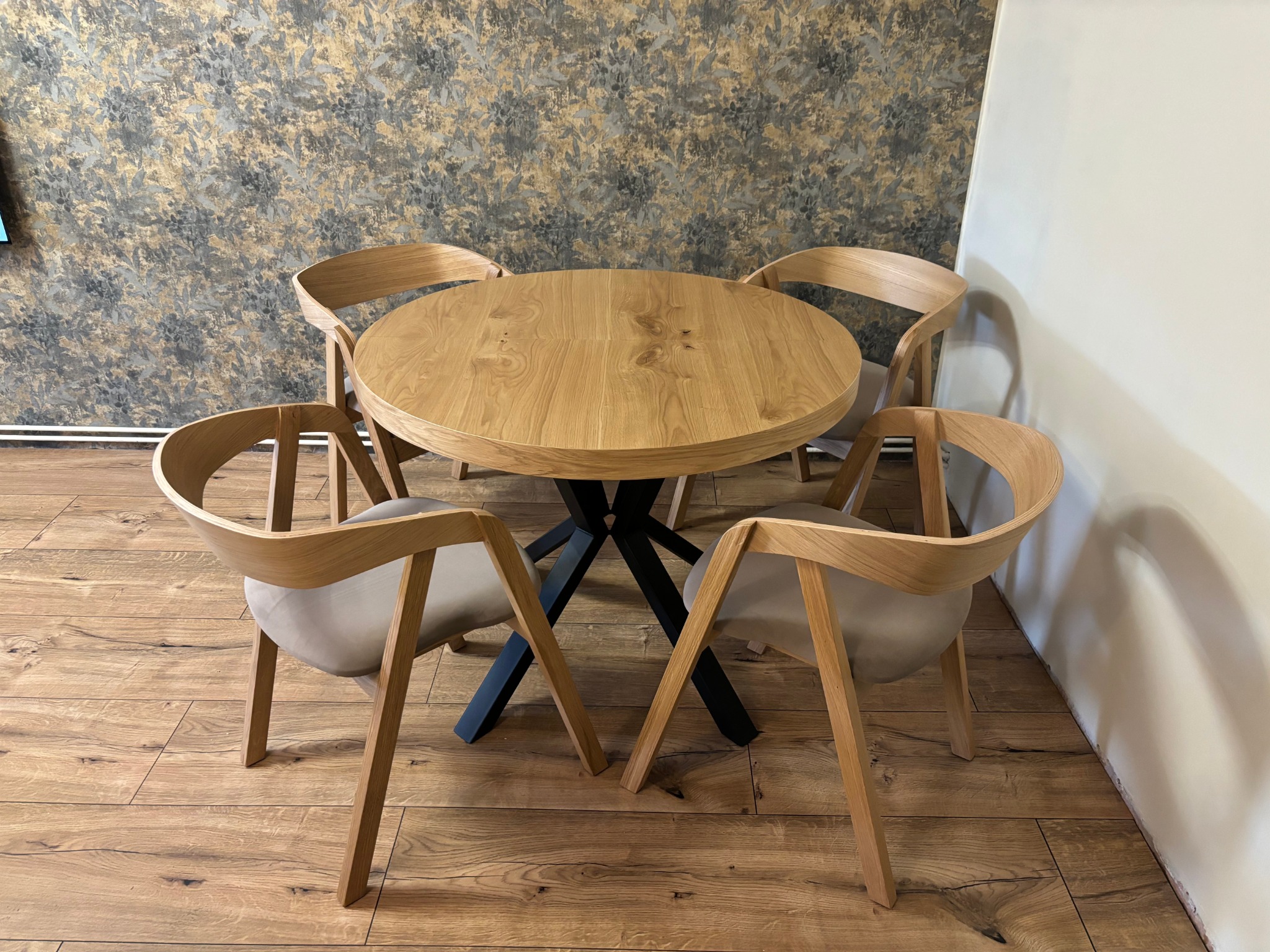 Drewniany okrągły stół z czarną podstawą i czterema krzesłami w jasnym drewnie, na tle wzorzystej tapety i drewnianej podłogi. Nowoczesny design.