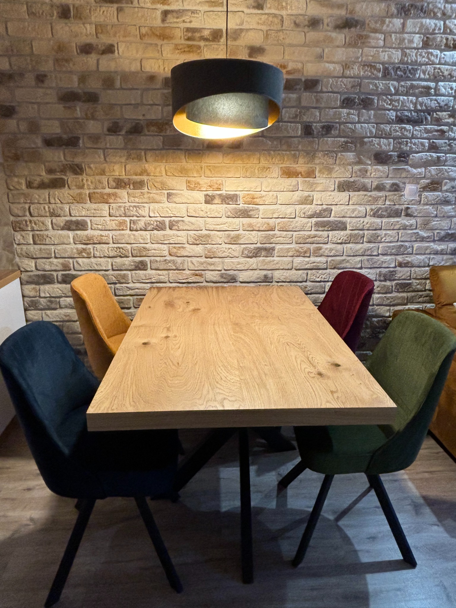 Drewniany stół na metalowych nogach otoczony krzesłami w różnych kolorach, na tle ściany z cegły i lampy wiszącej. Ustawienie mebli w jadalni.