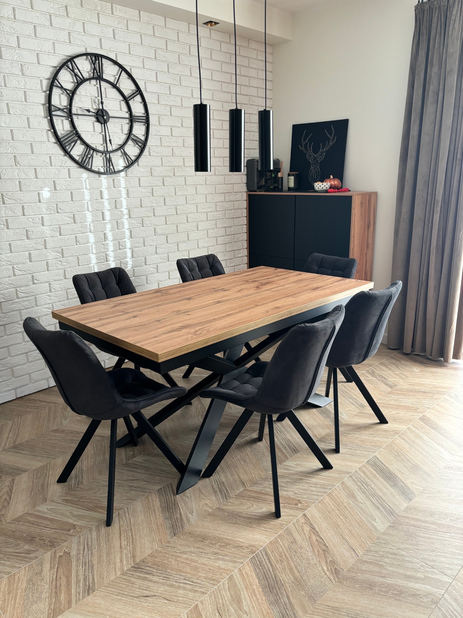 Jadalnia z drewnianym stołem na czarnych nogach, otoczonym sześcioma grafitowymi krzesłami. W tle biała ceglana ściana z zegarem i czarna komoda z dekoracjami.