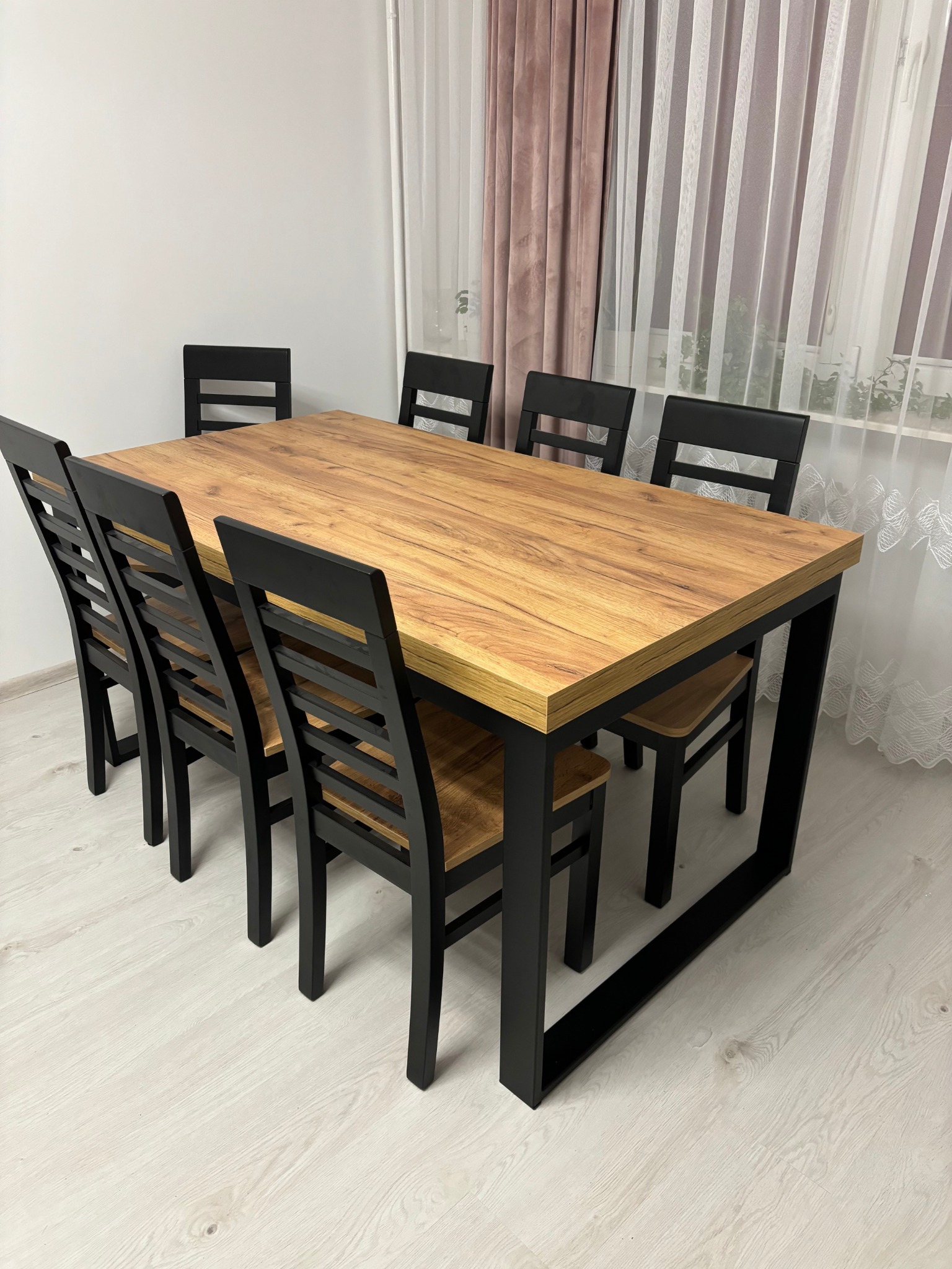 Stół z blatem imitującym drewno i czarnymi nogami oraz sześć krzeseł w pokoju z zasłonami. Zestaw mebli do jadalni.