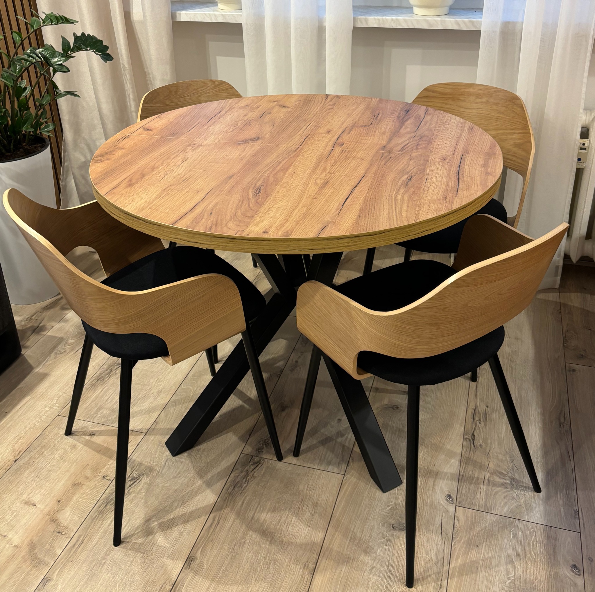 Okrągły stół z drewnianym blatem i czarną, metalową podstawą otoczony czterema krzesłami z drewnianymi oparciami i czarnymi siedziskami. W tle zasłony i roślina.