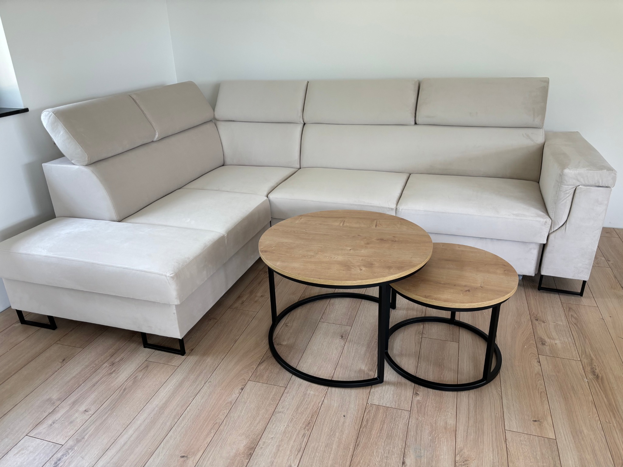 Jasny, narożny wypoczynek z regulowanymi zagłówkami i stoliki kawowe na drewnianej podłodze. Minimalistyczny design, neutralna kolorystyka.