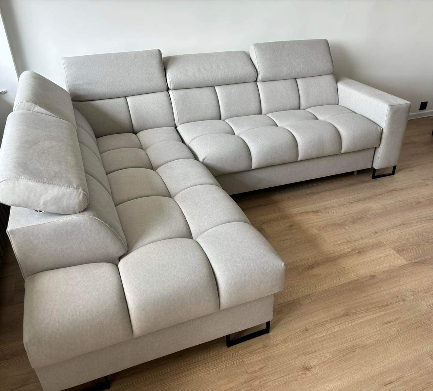 Jasnoszary narożnik z pikowanymi siedziskami i minimalistycznymi nóżkami na drewnianej podłodze, ustawiony pod białą ścianą. Nowoczesny design.