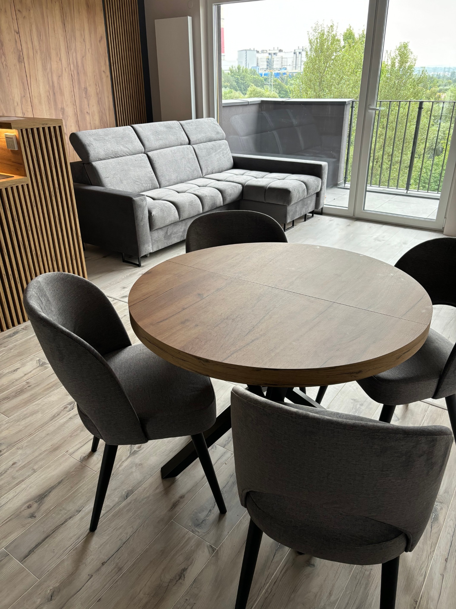 Salon z szarą sofą narożną, okrągłym drewnianym stołem i krzesłami na balkonie z widokiem na zieleń. Drewniana lamela na ścianie dodaje ciepła.