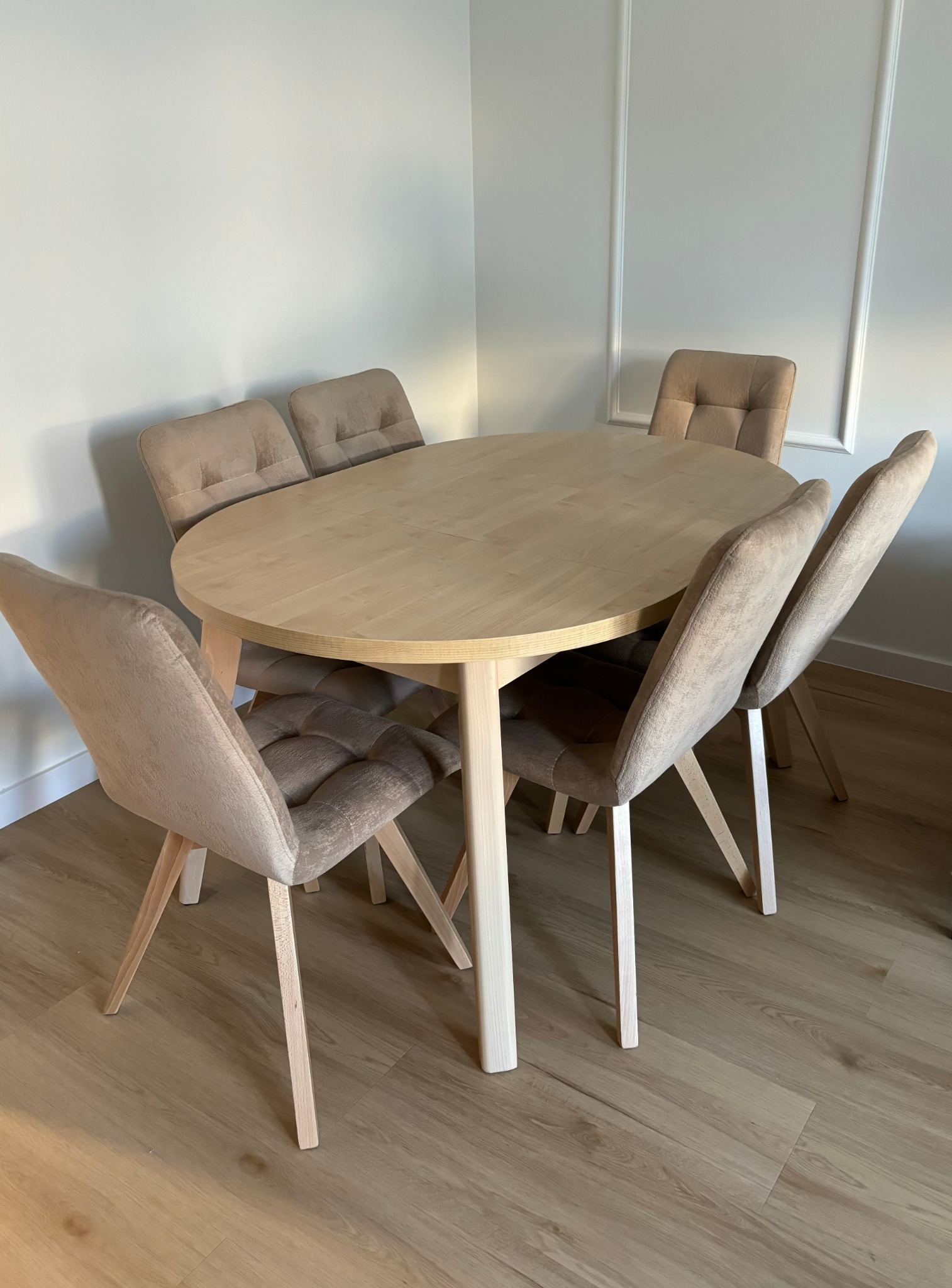 Jasny, owalny stół z sześcioma krzesłami w beżowych odcieniach, ustawiony w rogu pokoju z dekoracyjnymi listwami przypodłogowymi. Minimalistyczny design.