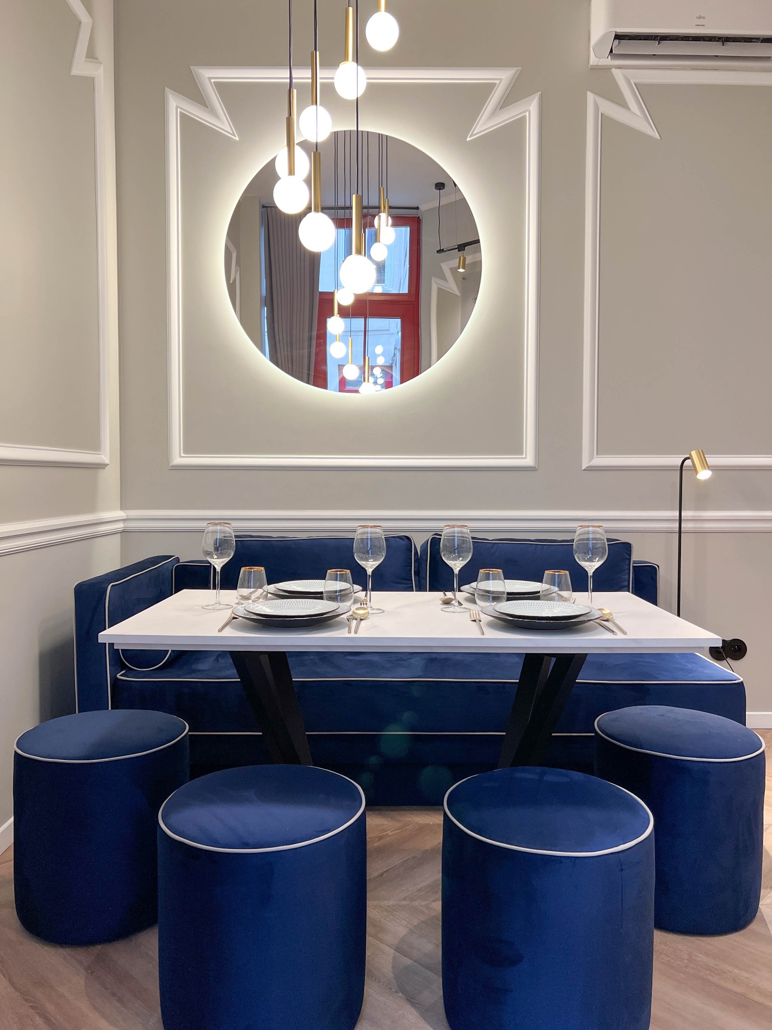Elegancka jadalnia z niebieską sofą zamiast krzeseł, białym stołem i okrągłym lustrem z podświetleniem. Cztery pufy w kolorze sofy dopełniają aranżację.