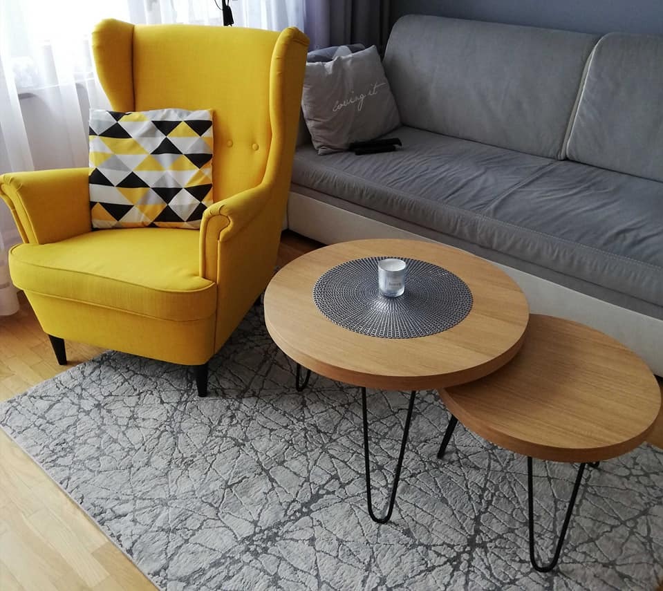 Żółty fotel typu uszak z poduszką w geometryczne wzory, stoliki kawowe z drewnianymi blatami i metalowymi nogami na tle szarej sofy i dywanu.