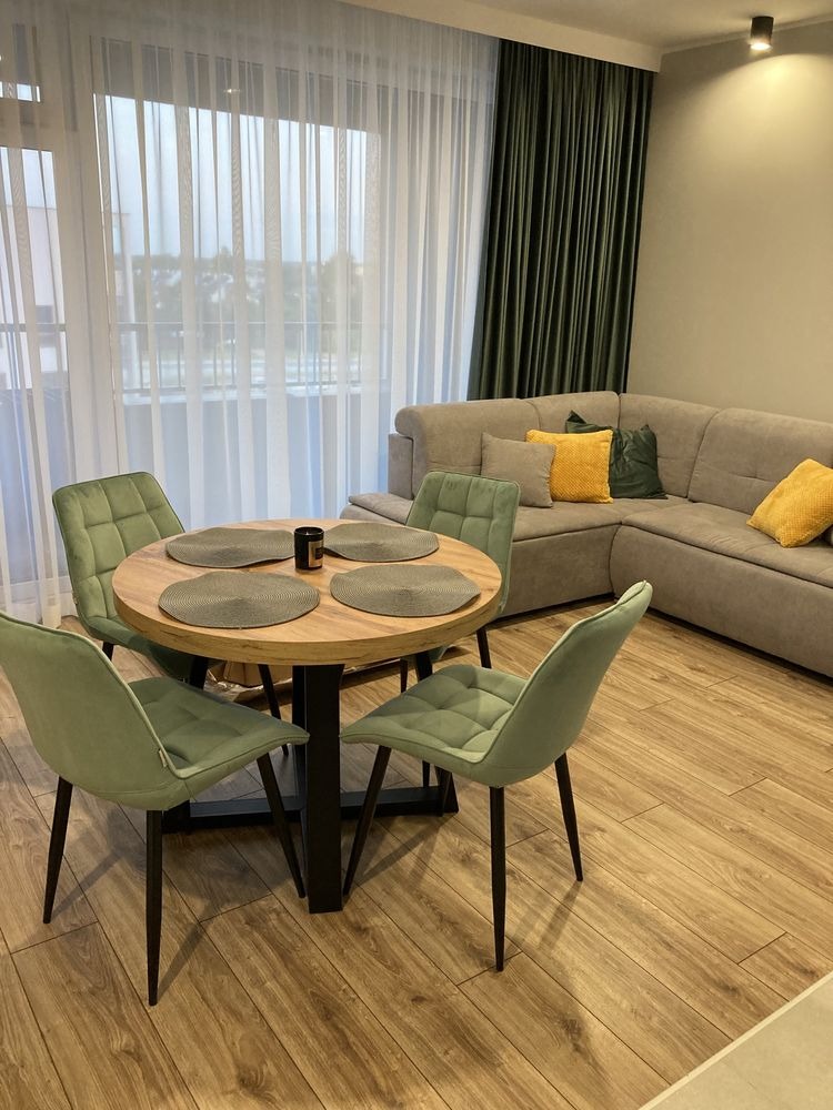 Jasne wnętrze z okrągłym stołem, zielonymi krzesłami i narożnikiem. Parkiet imitujący drewno. Stonowana kolorystyka z akcentami zieleni i żółci. Minimalistyczny styl.