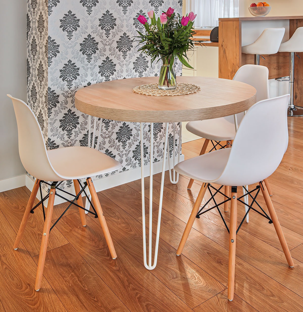 Okrągły stół z blatem z jasnego drewna i białymi nogami w stylu hairpin, otoczony krzesłami z drewnianymi nogami. Na stole bukiet różowych tulipanów.