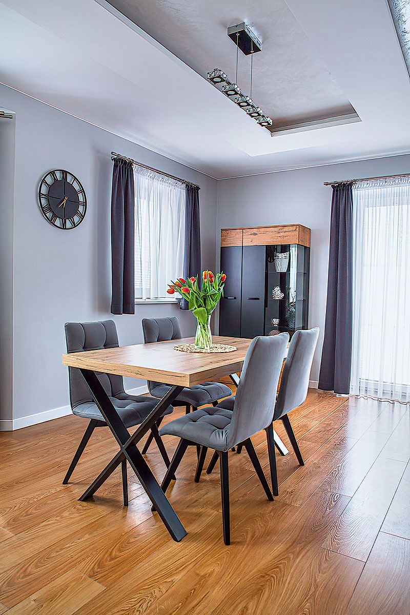 Nowoczesna jadalnia: drewniany stół z bukietem tulipanów, szare krzesła na metalowych nogach, szafka z przeszklonym frontem i ozdobny zegar na szarej ścianie.