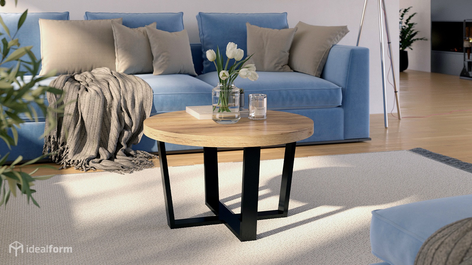 Okrągły stolik kawowy z drewnianym blatem i czarnymi nogami na tle błękitnej sofy z poduszkami i dywanu. Na stoliku wazon z białymi tulipanami i świeca.
