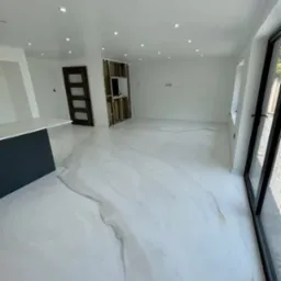 Jasne wnętrze z białą posadzką o wzorze marmuru. Minimalistyczny design, duże okno, widoczne drzwi i fragment zabudowy kuchennej. Nowoczesne oświetlenie sufitowe.