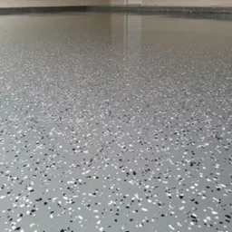 Szara posadzka żywiczna z czarnymi i białymi płatkami, gładka powierzchnia, widoczne odbicie światła, wnętrze garażu lub hali.