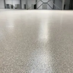 Jasnoszara, gładka posadzka w hali przemysłowej, z widocznymi metalowymi słupami konstrukcyjnymi w tle. Delikatny połysk na powierzchni podłogi.