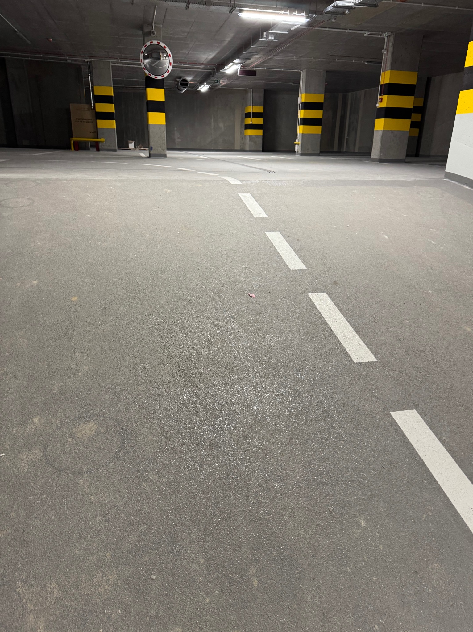 Szara posadzka w garażu podziemnym z białymi liniami, słupy oznaczone czarno-żółtymi pasami. Widok na przestrzeń parkingu z oświetleniem sufitowym.