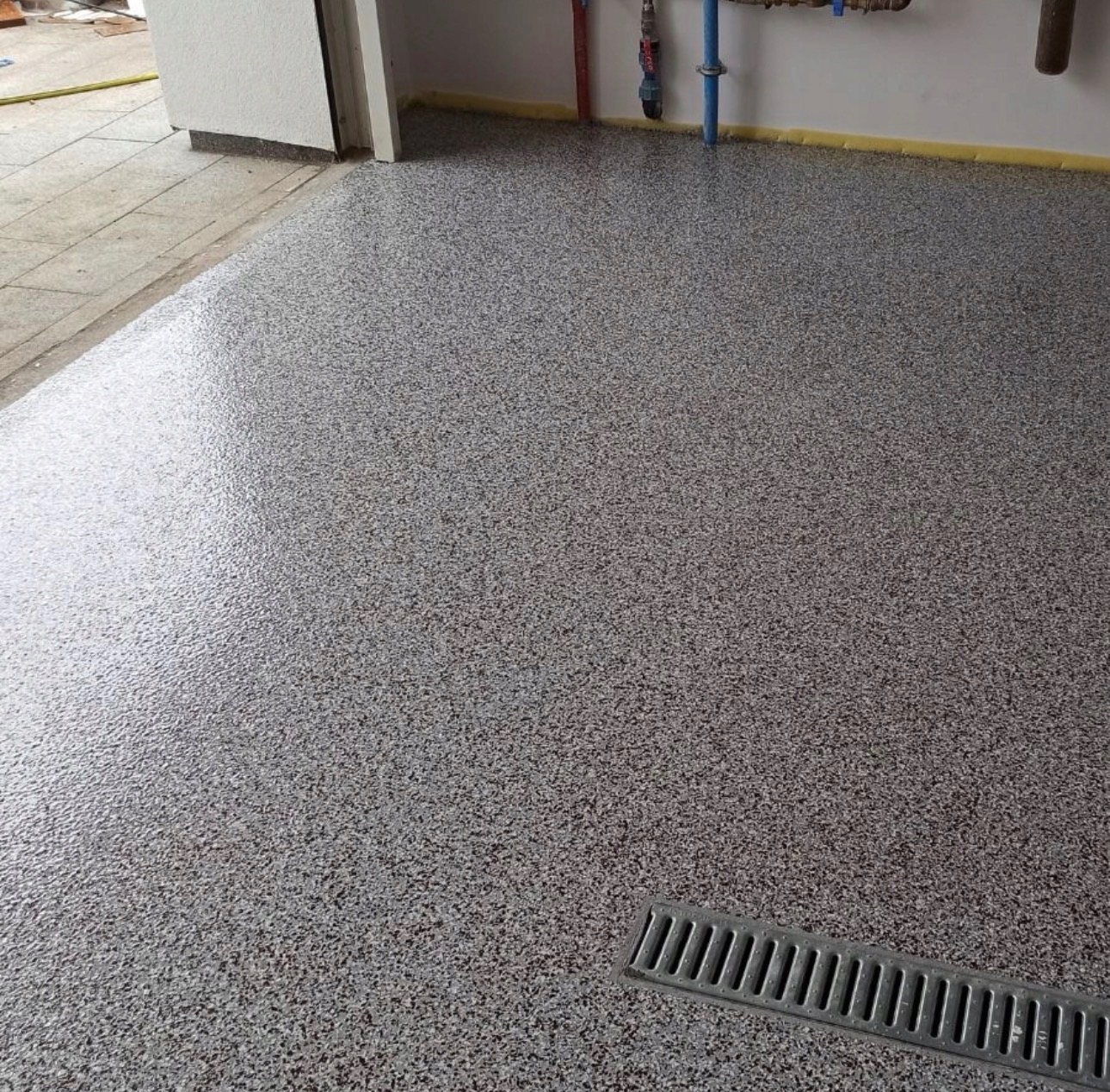 Szara, błyszcząca posadzka z drobnymi plamkami, wykończona przy ścianie żółtą taśmą maskującą. W rogu widoczny odpływ liniowy.