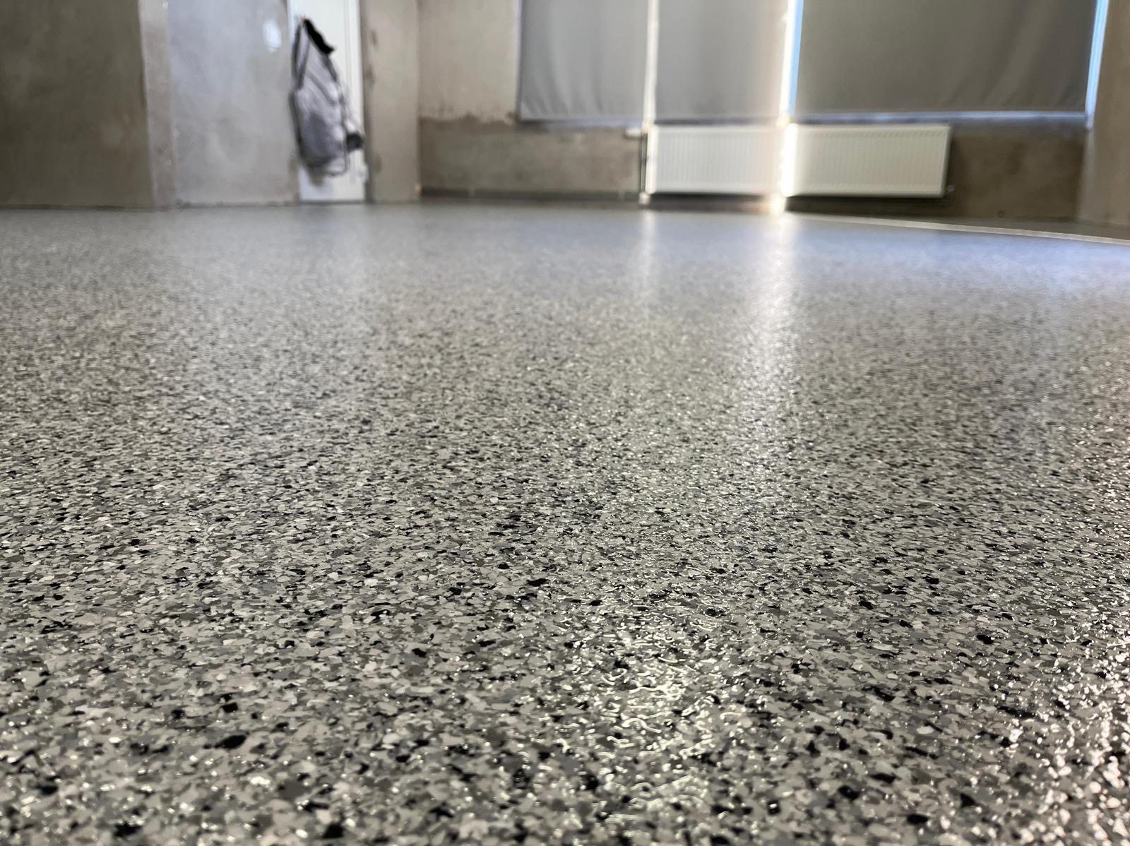 Błyszcząca, szara posadzka z drobnymi, czarnymi elementami w surowym wnętrzu z grzejnikiem i oknami w tle. Nowoczesne wykończenie podłogi.