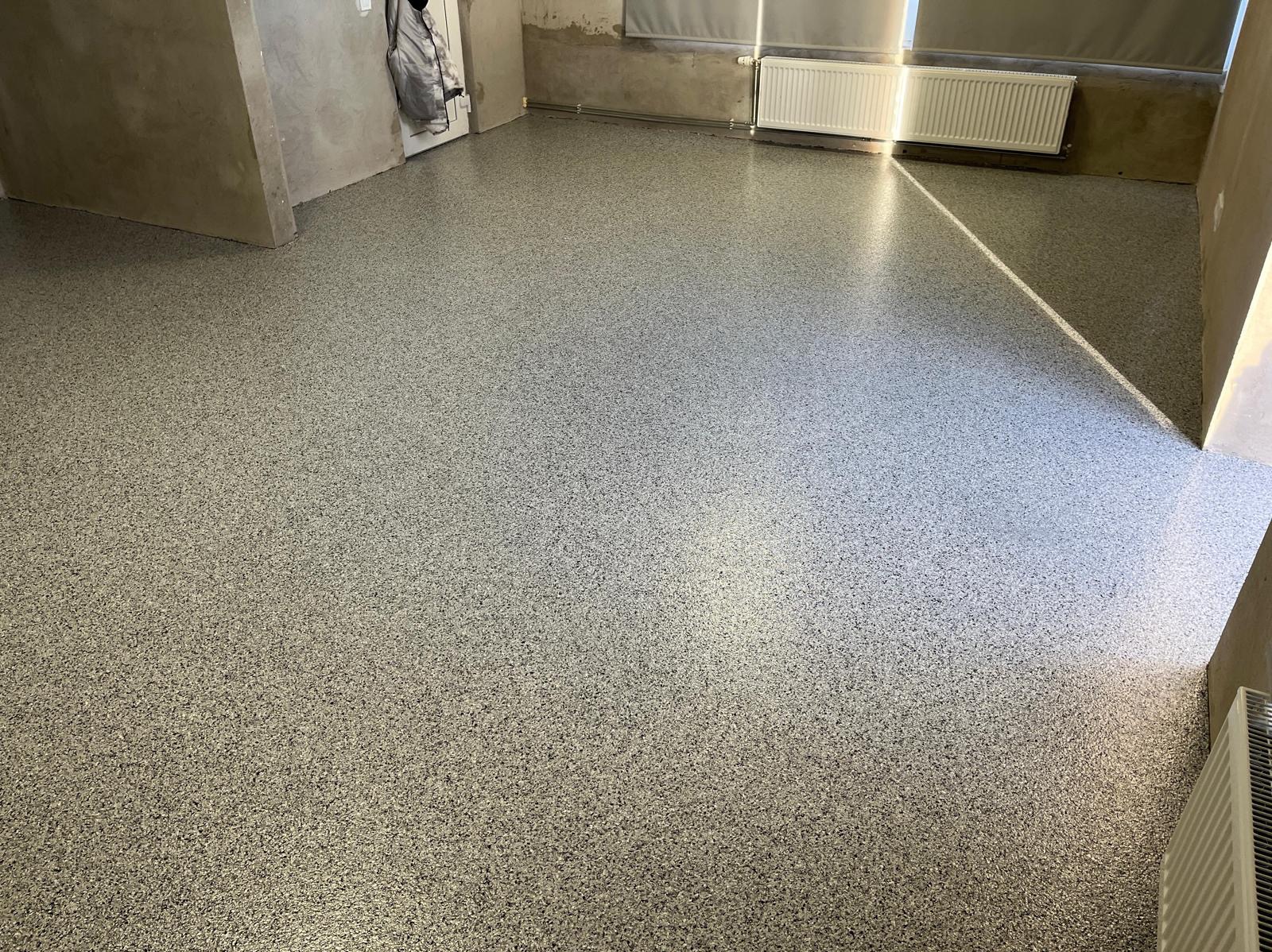 Jasnoszara, błyszcząca podłoga z żywicy w surowym wnętrzu, z widocznym grzejnikiem i fragmentem ściany w stanie deweloperskim. Widoczne promienie słoneczne.