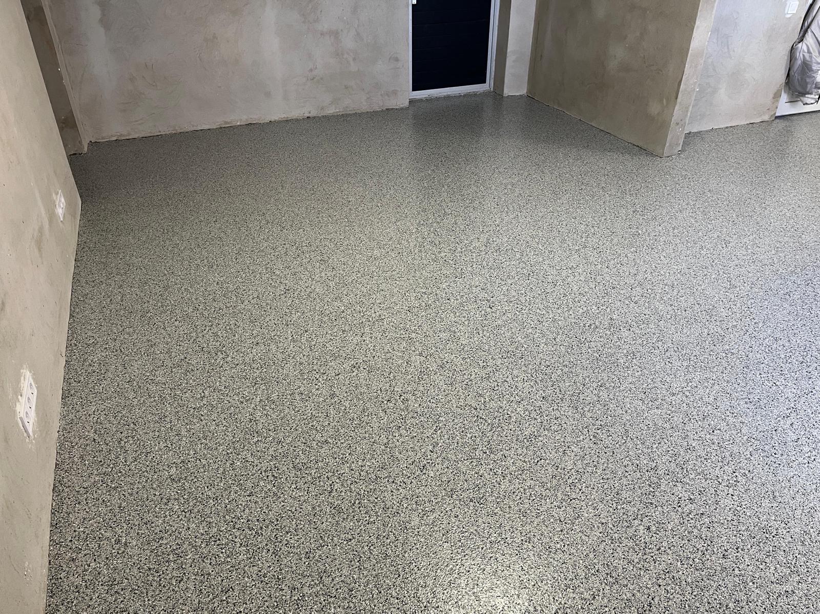 Błyszcząca, szara posadzka żywiczna z drobnym wzorem w surowym wnętrzu. Widoczne ściany w stanie deweloperskim i czarne drzwi w tle. Nowoczesne wykończenie podłogi.