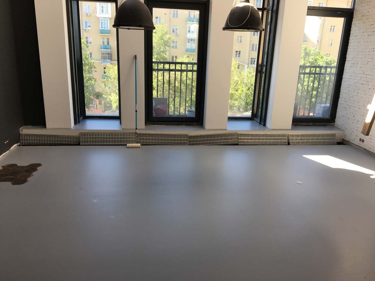 Szara, gładka posadzka w jasnym wnętrzu z dużymi oknami. Widoczny wałek malarski i fragment starej posadzki. Dwa wiszące, czarne klosze lamp.